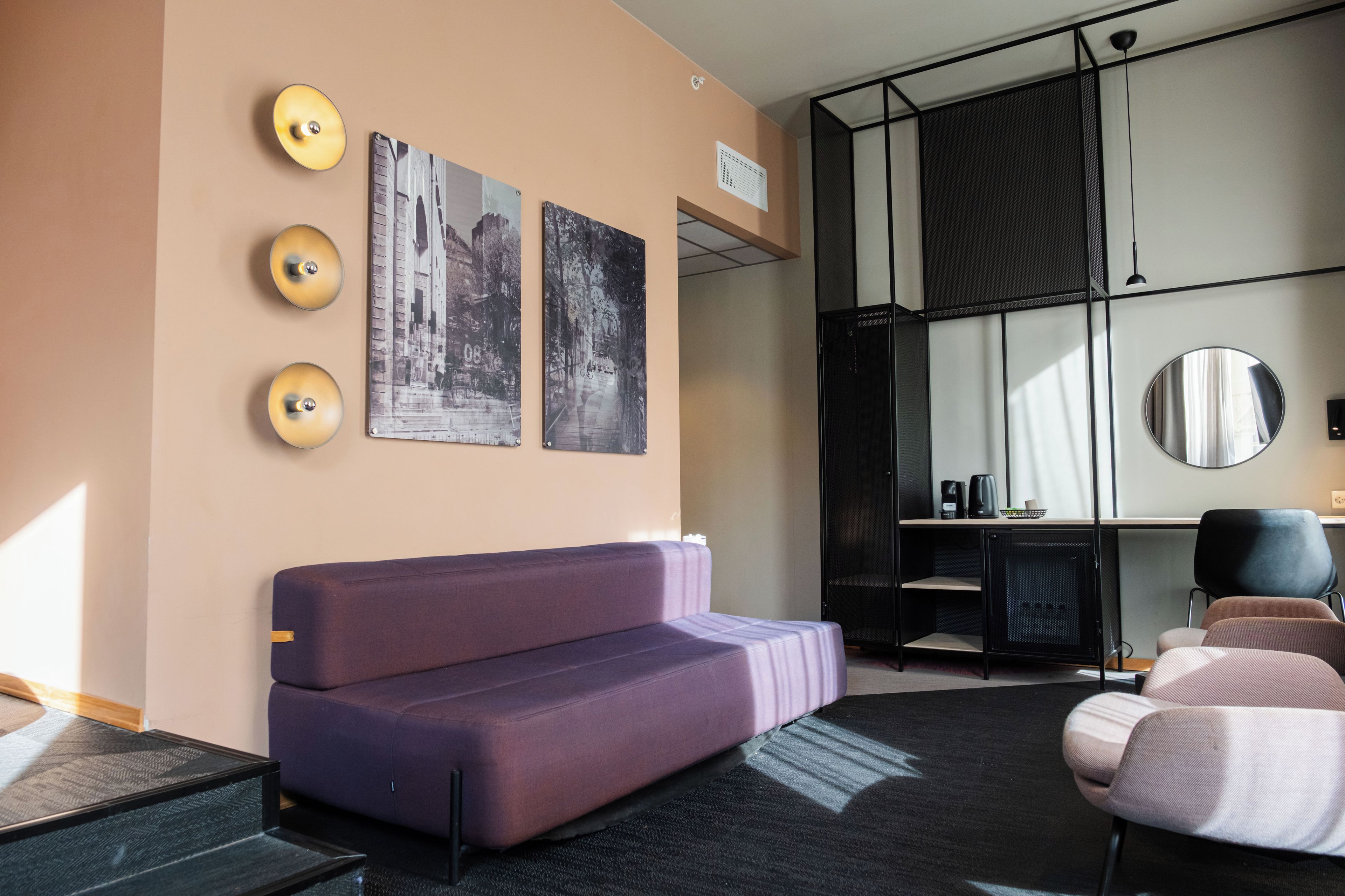 A modern hotel room with a purple sofa, a desk with a computer, a large mirror, and two wall-mounted artworks.