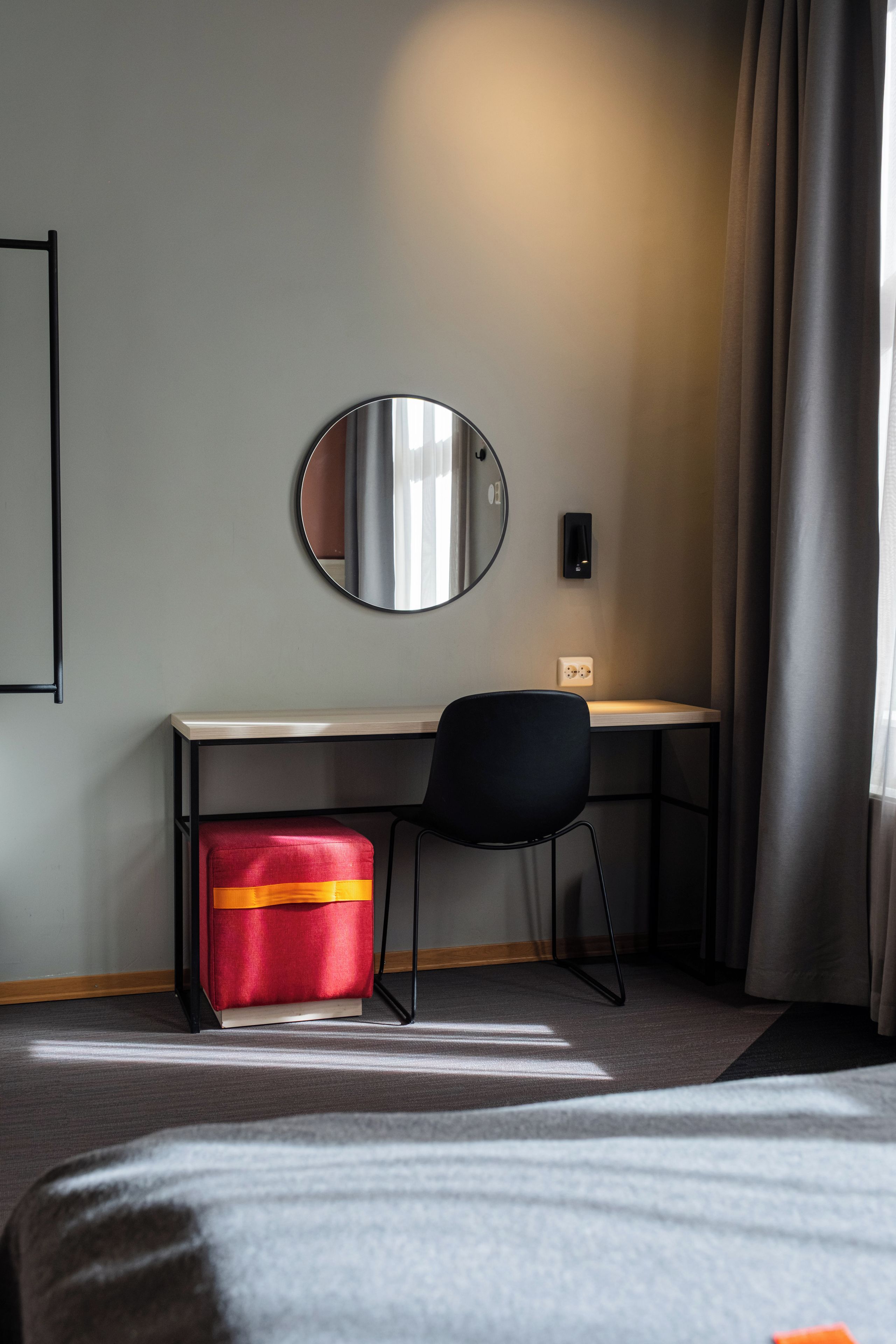 Une chambre d’hôtel avec un lit, un bureau, une chaise, une valise rouge, un miroir rond et une lampe fixée au mur.