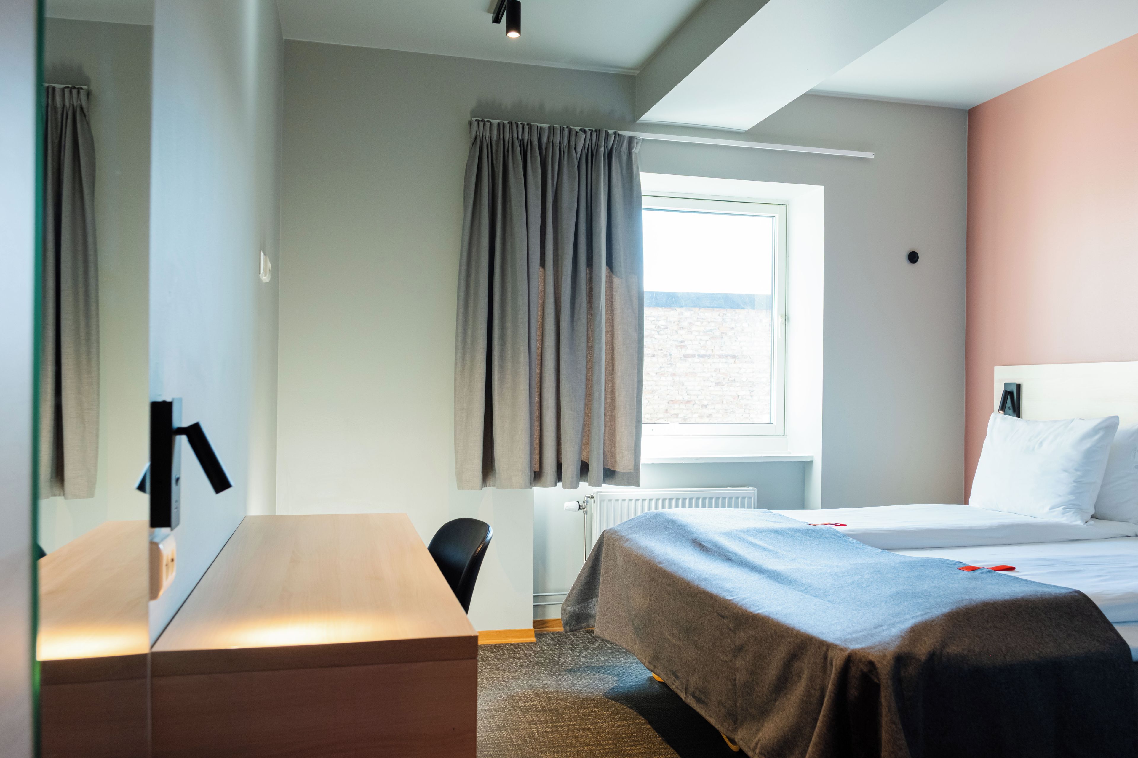 A hotel room with a bed, a desk, a chair, a window with curtains, and a radiator.