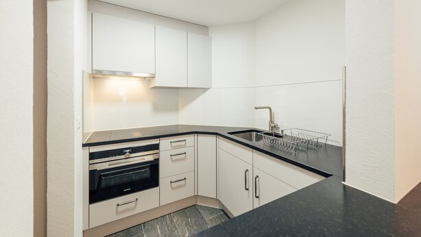 A compact kitchen with white cabinets, a black countertop, and a built-in oven. - Hotel Kruezli (Tujetsch)