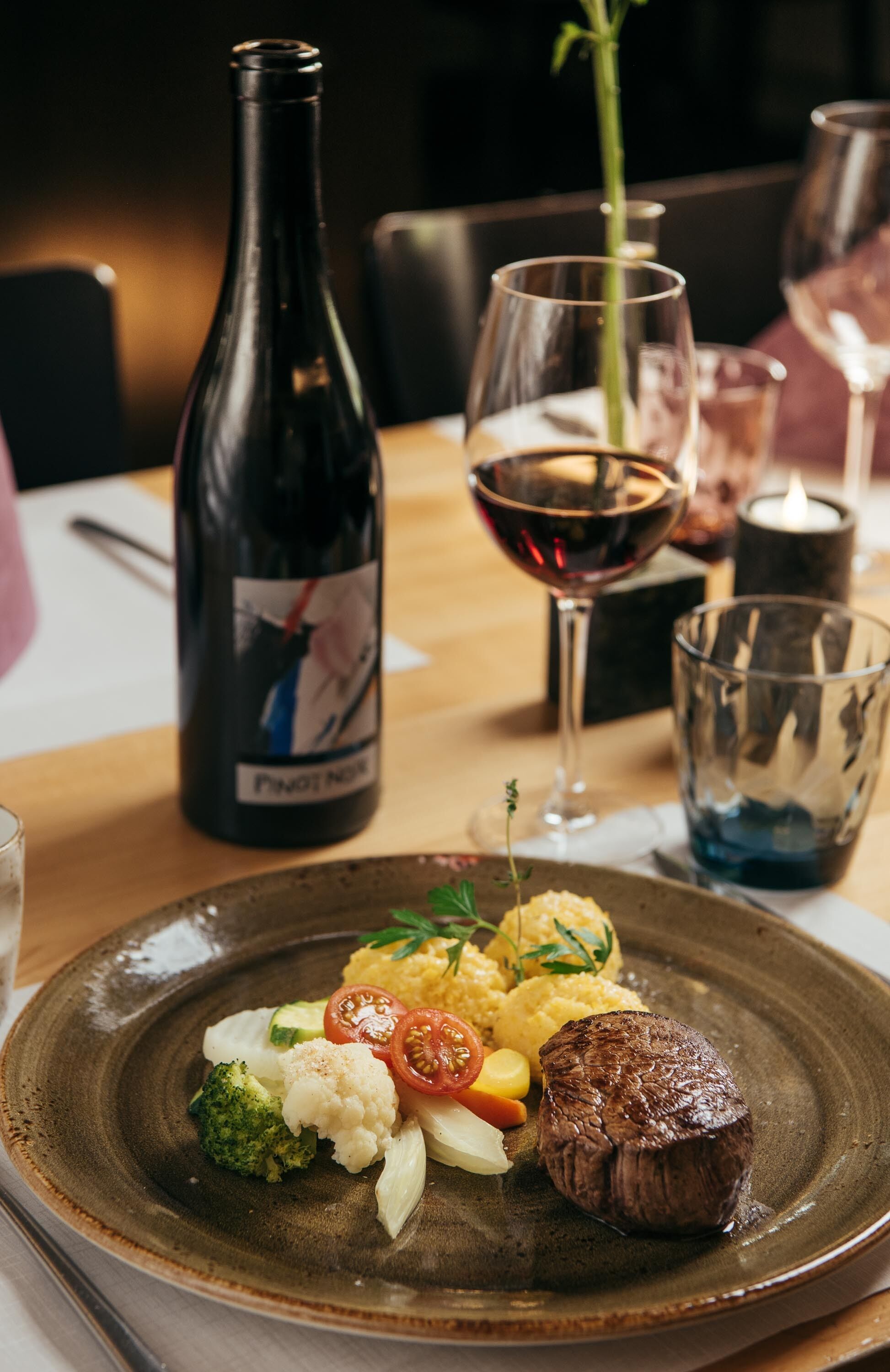 Een bord eten met een steak, groenten en puree, vergezeld van een fles wijn en een glas rode wijn.