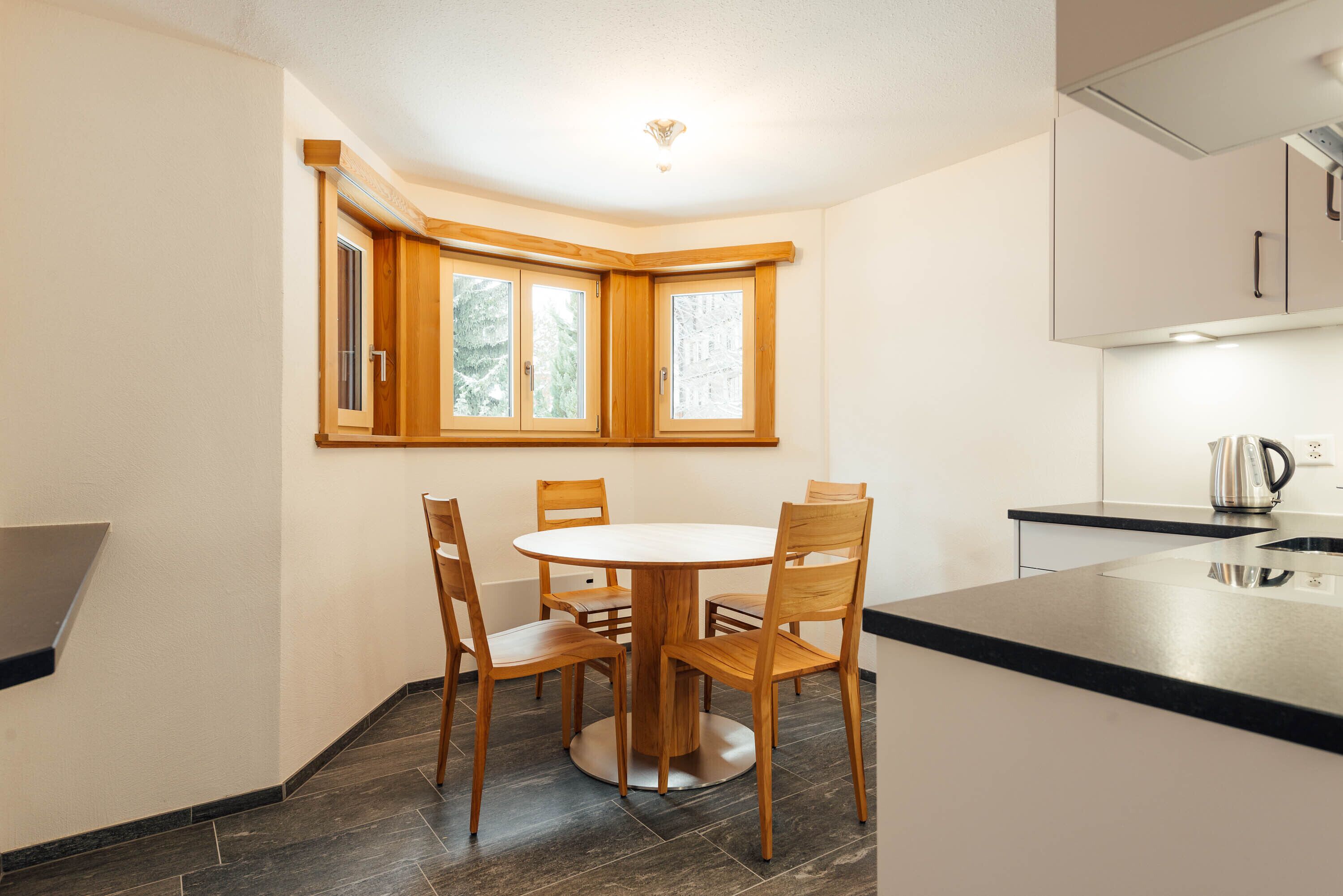 A corner kitchen with a round dining table and four chairs, a window with wooden frames, and a black countertop.
