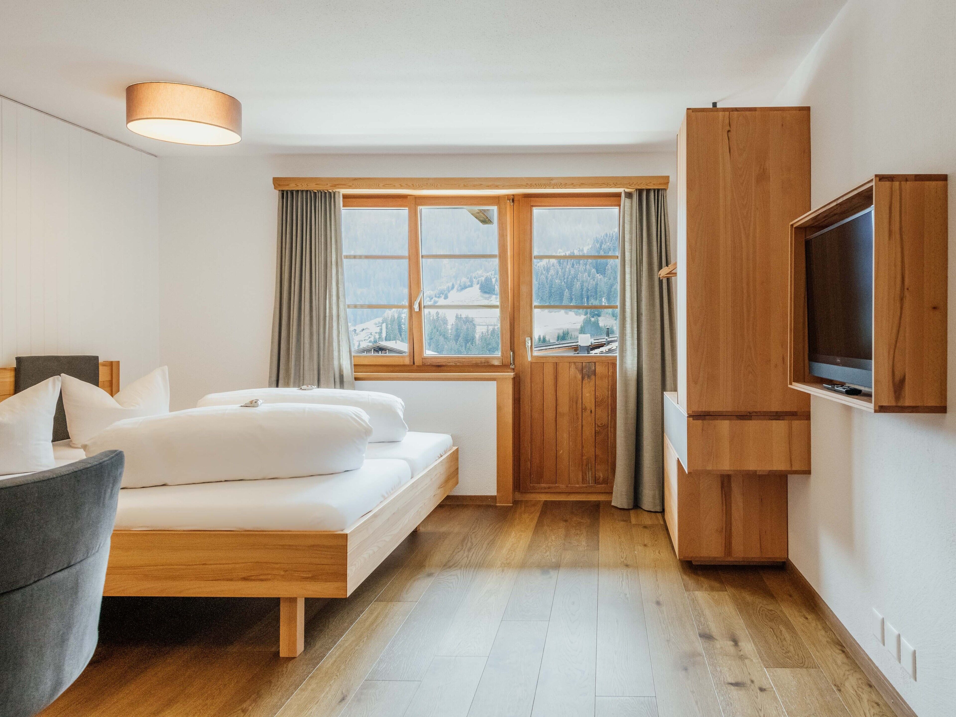 A modern bedroom with a wooden bed, a grey chair, a television, and a window with a view of snow-covered mountains.