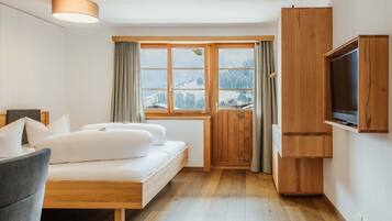 A modern bedroom with a wooden bed, a grey chair, a television, and a window with a view of snow-covered mountains.
