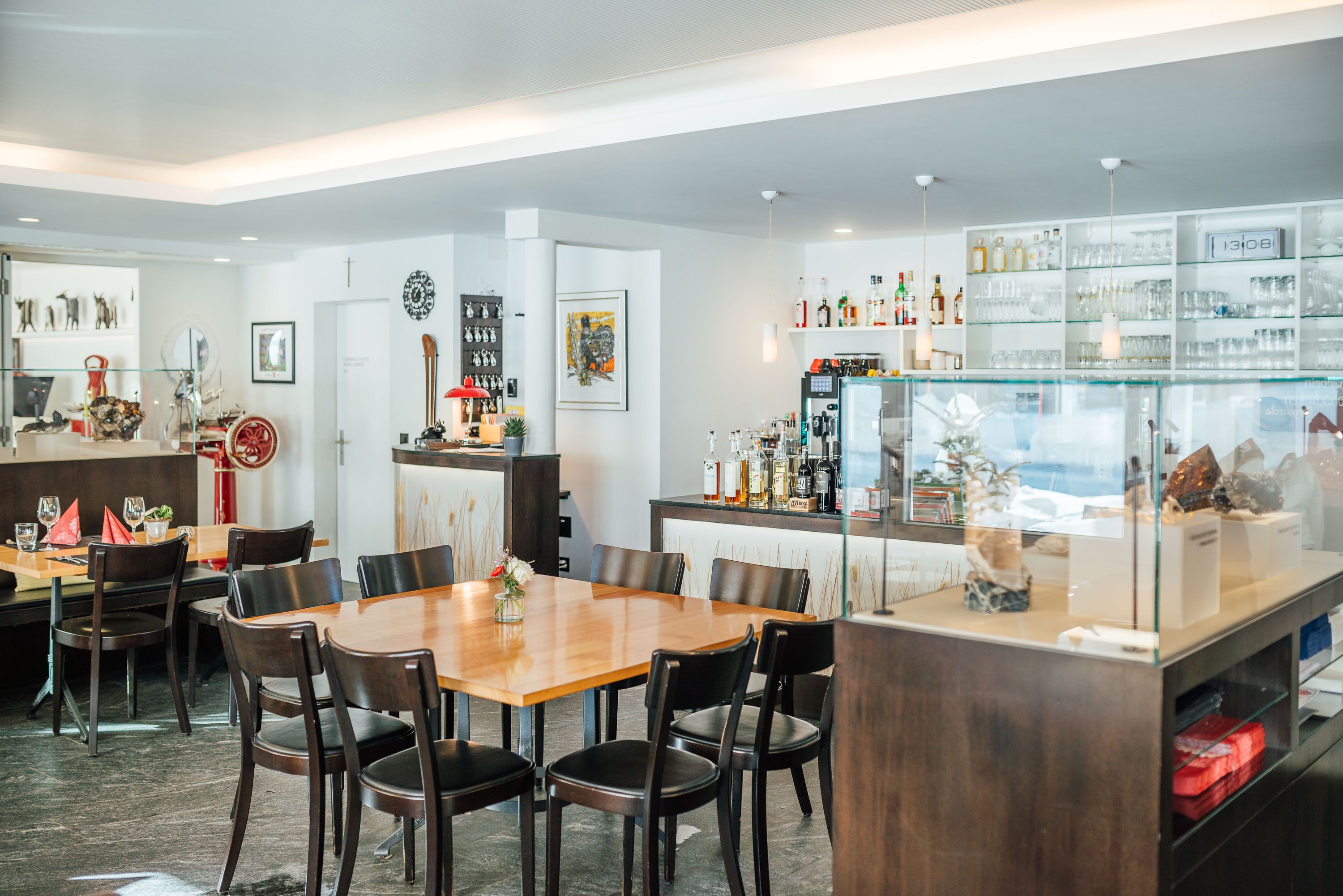 Un comedor moderno con una mesa y sillas de madera, una zona de cocina con estanterías de vidrio y diversos objetos decorativos.