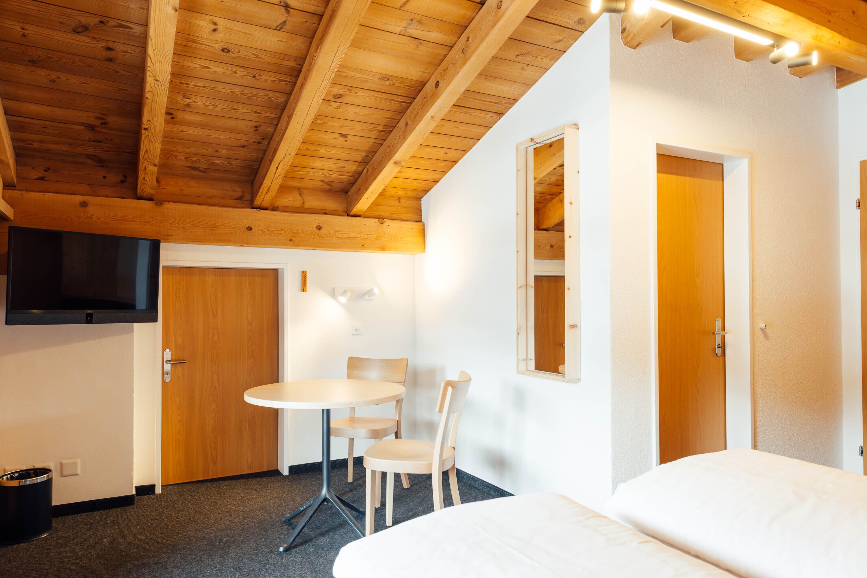 Habitación de hotel con techo de madera, una cama, una pequeña mesa redonda con dos sillas, un televisor montado en la pared y una puerta.