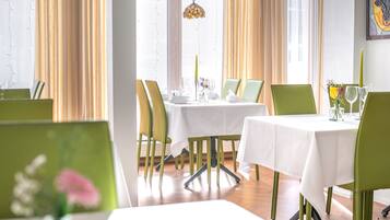 Uma sala de jantar com mesas postas para quatro pessoas, cadeiras verdes e toalha de mesa branca.