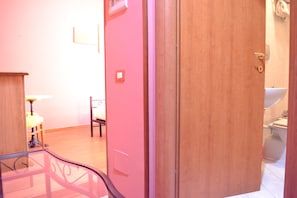 Une pièce avec une porte en bois, une petite table avec une lampe, une chaise et une salle de bain visible à travers la porte.