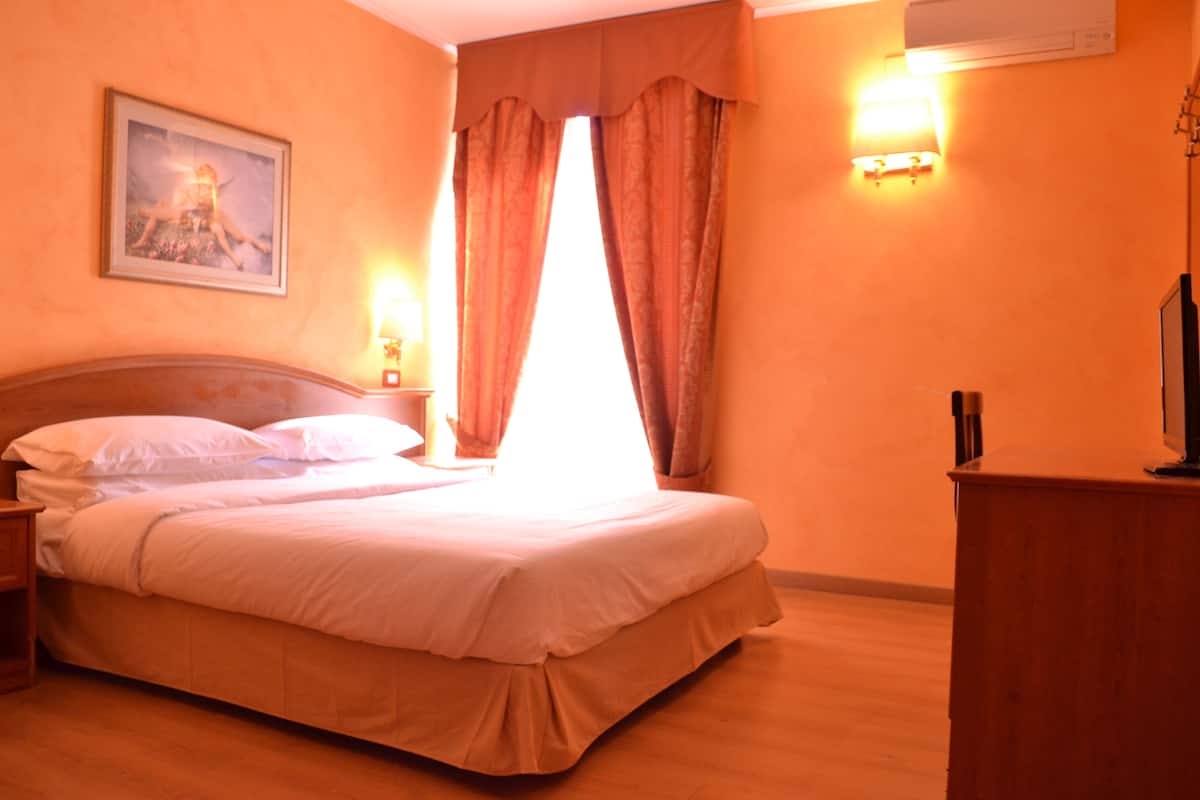 Una habitación de hotel con una cama, una mesita de noche, una lámpara, un cuadro en la pared y una ventana con cortinas.