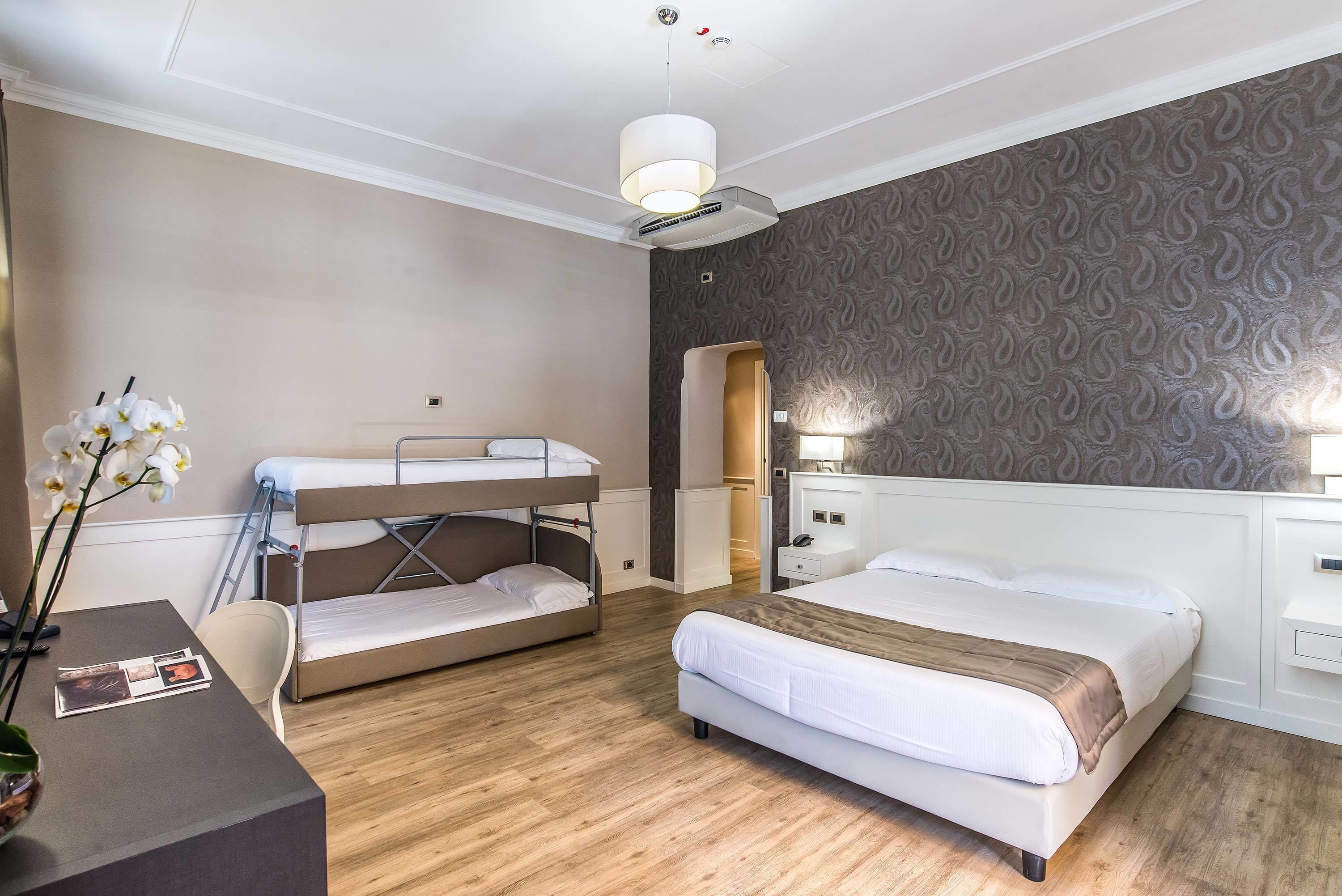A hotel room with bunk beds, a desk with a vase of flowers, a bedside table, and a patterned wall.