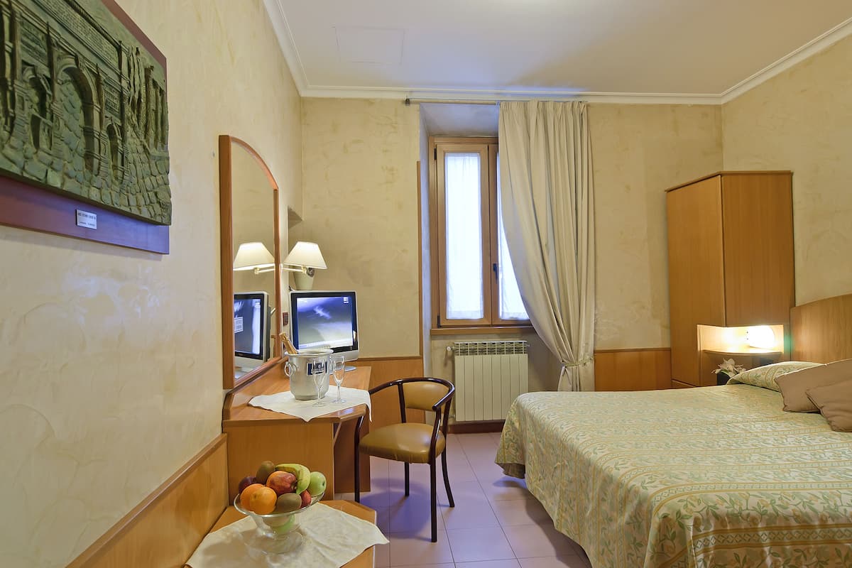 Habitación de hotel con una cama, un escritorio con computadora, una silla, un frutero y un cuadro en la pared.