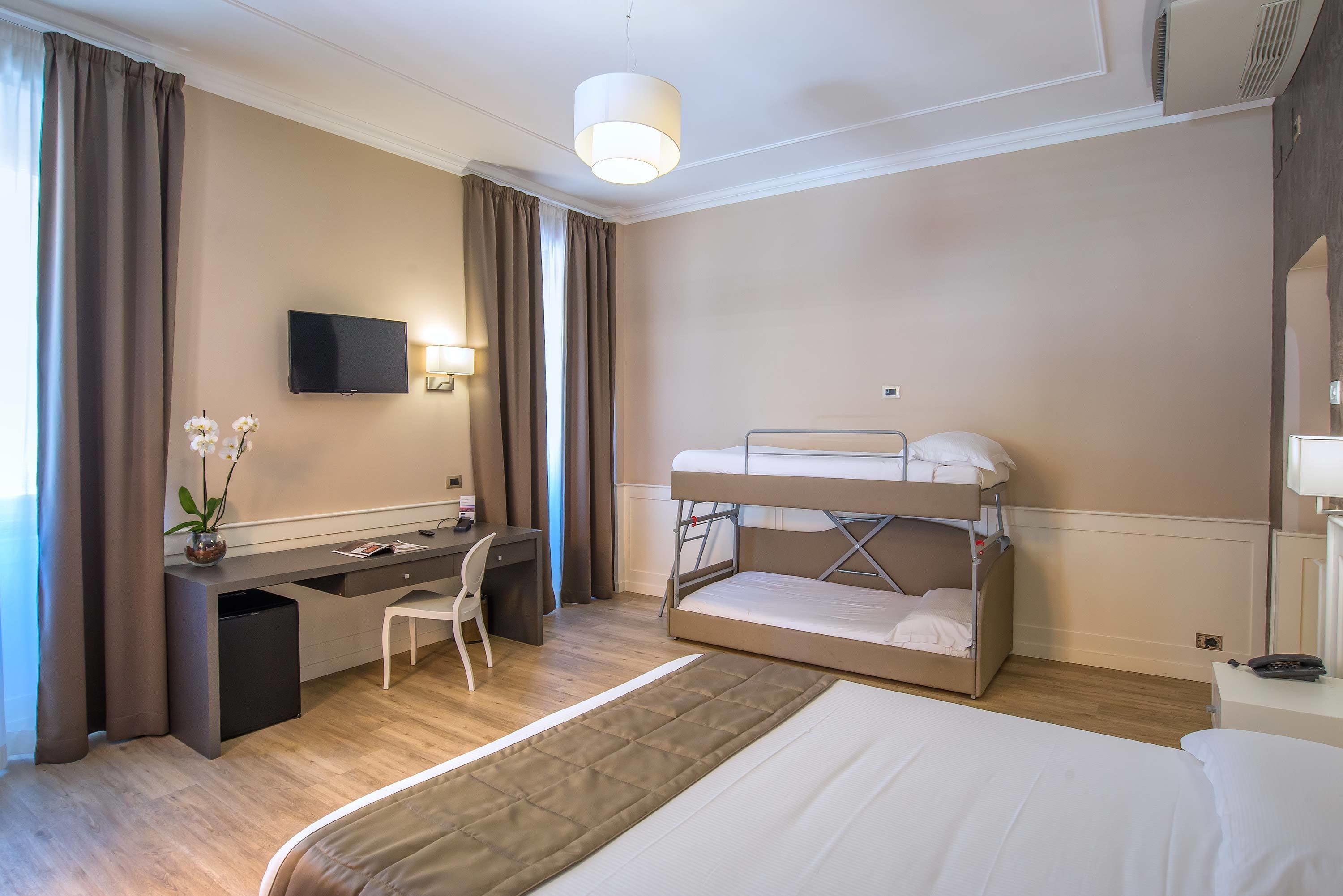 A hotel room with a bunk bed, a desk with a chair, a television, and a bedside table with a lamp.