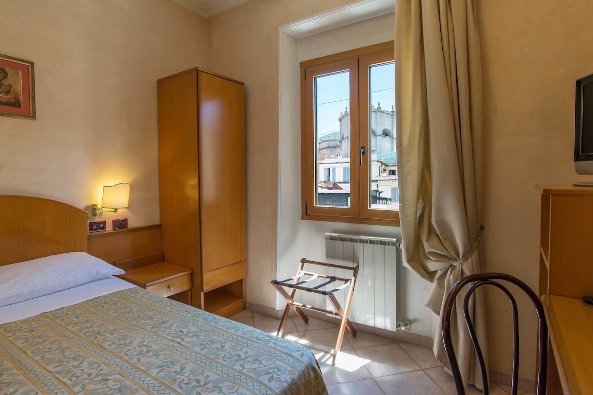 Habitación de hotel con cama, armario de madera, radiador, ventana con vistas a edificios y una silla.