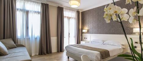 A modern hotel room with a large bed, a sofa, a desk, and a potted orchid.