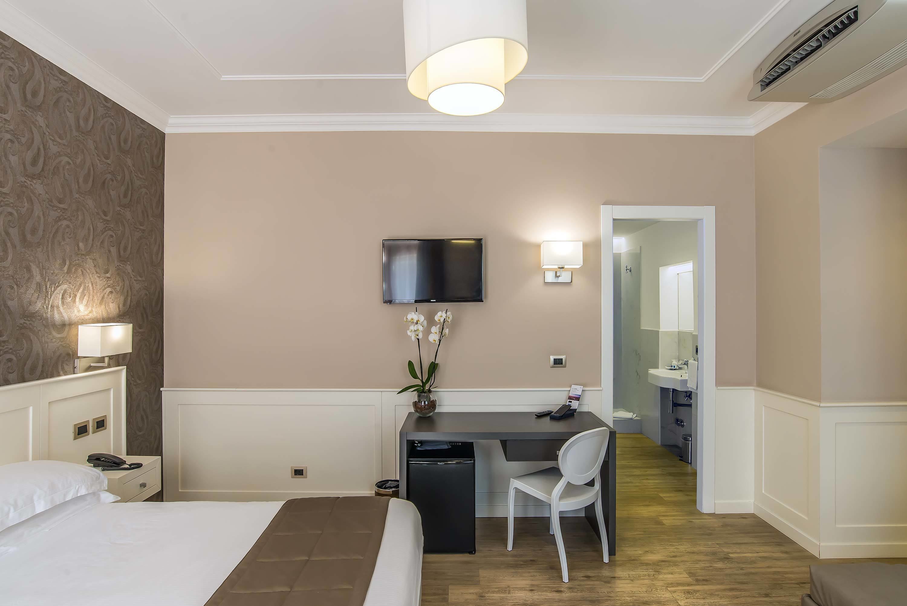 A hotel room with a bed, desk, chair, TV, and a small desk lamp.