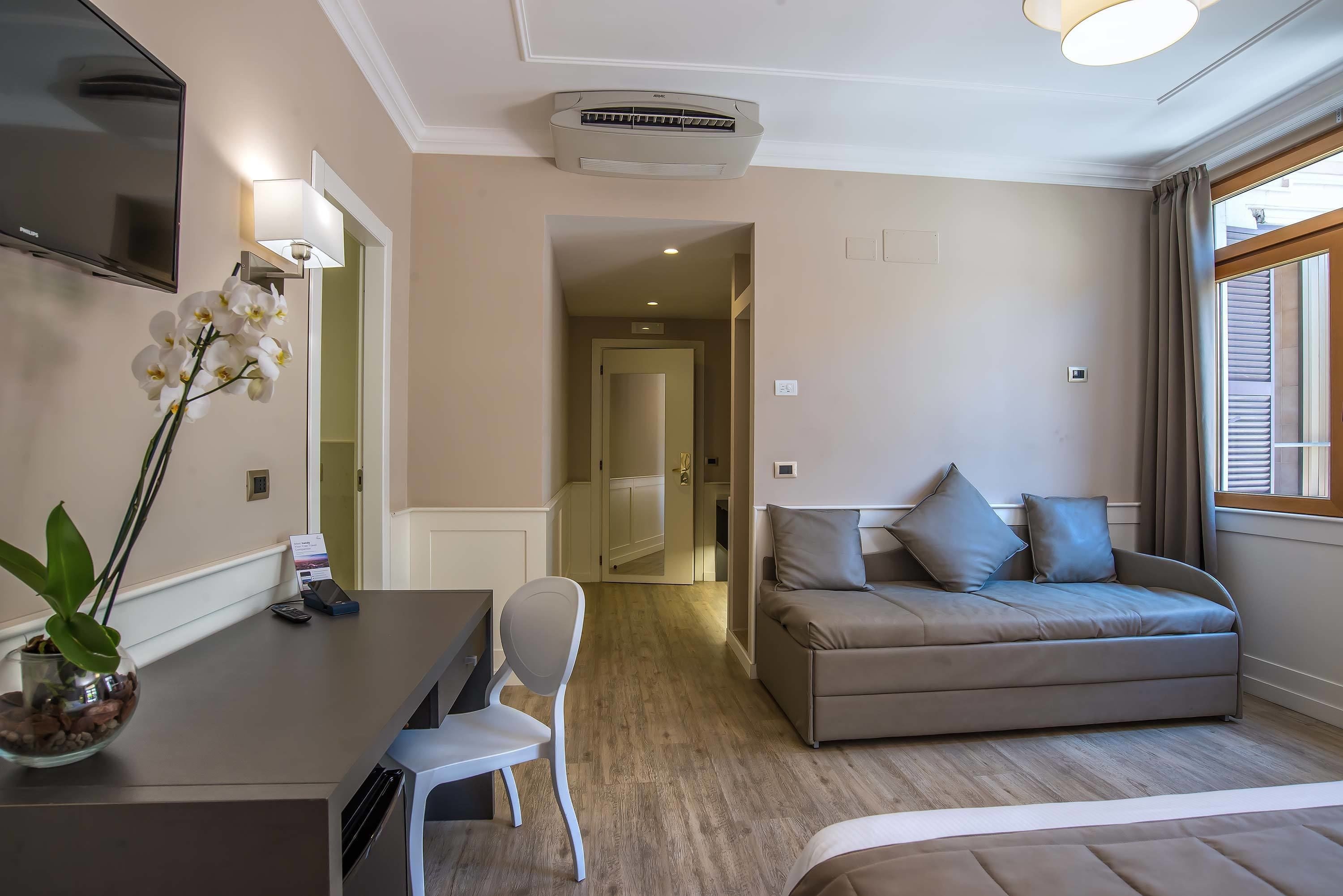 A hotel room with a sofa, a desk with a chair, a TV, and a potted plant.