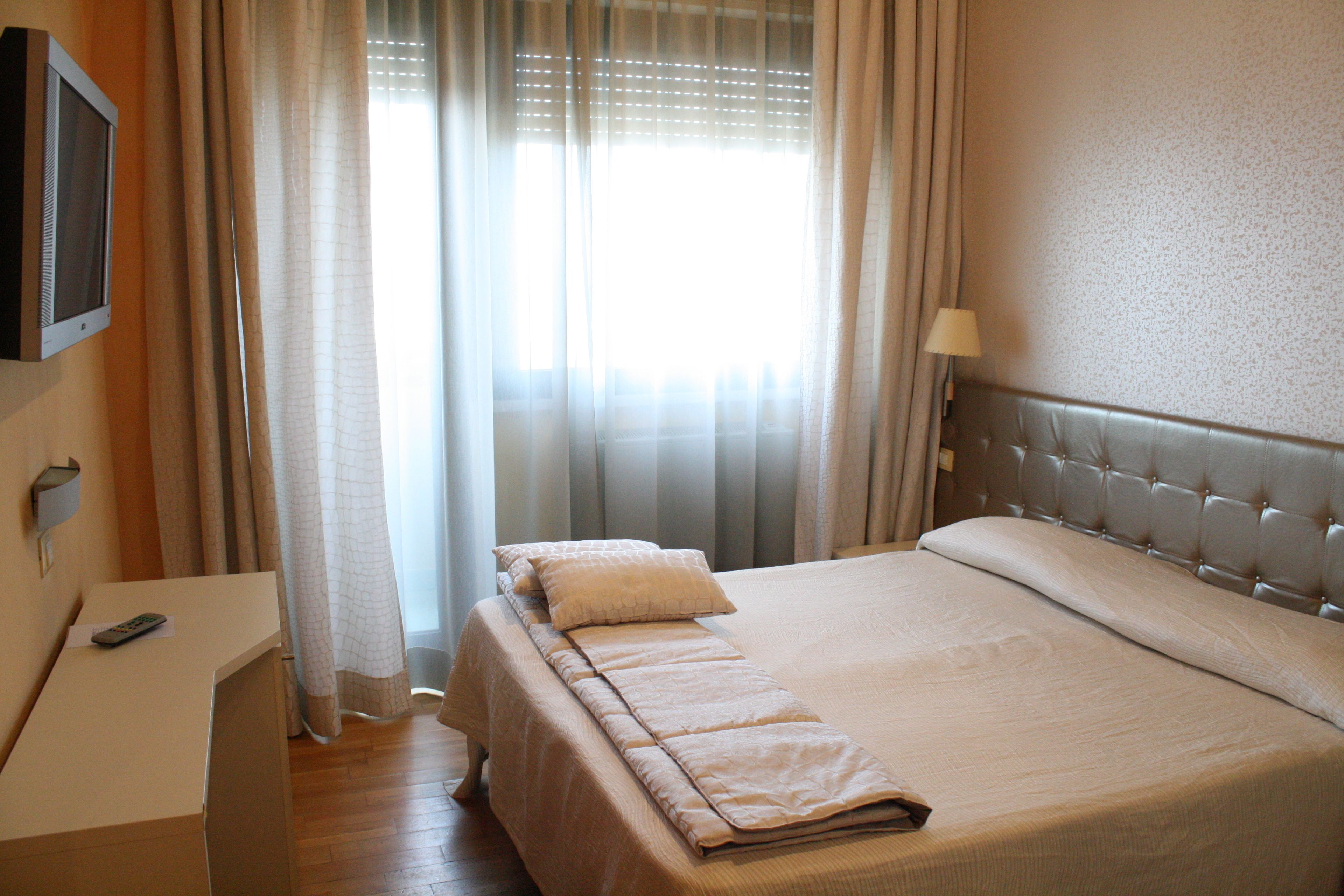 A hotel room with a bed, a bedside table, a remote control, and a window with curtains.