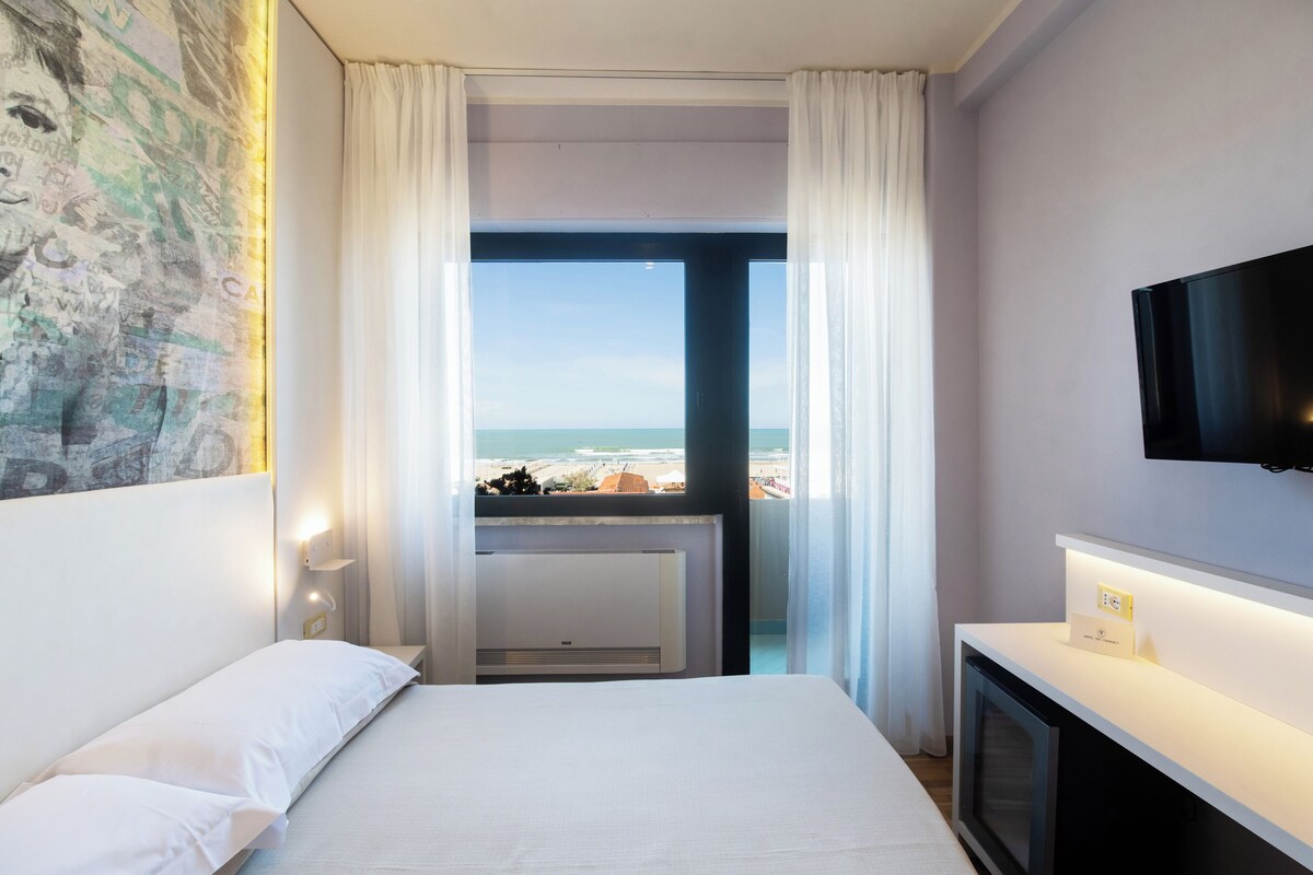 Habitación de hotel con cama, televisión, vista a la playa y ventana con cortinas.