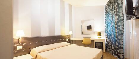 Una habitación de hotel con cama, mesitas de noche, escritorio, espejo, televisión y cortina estampada.