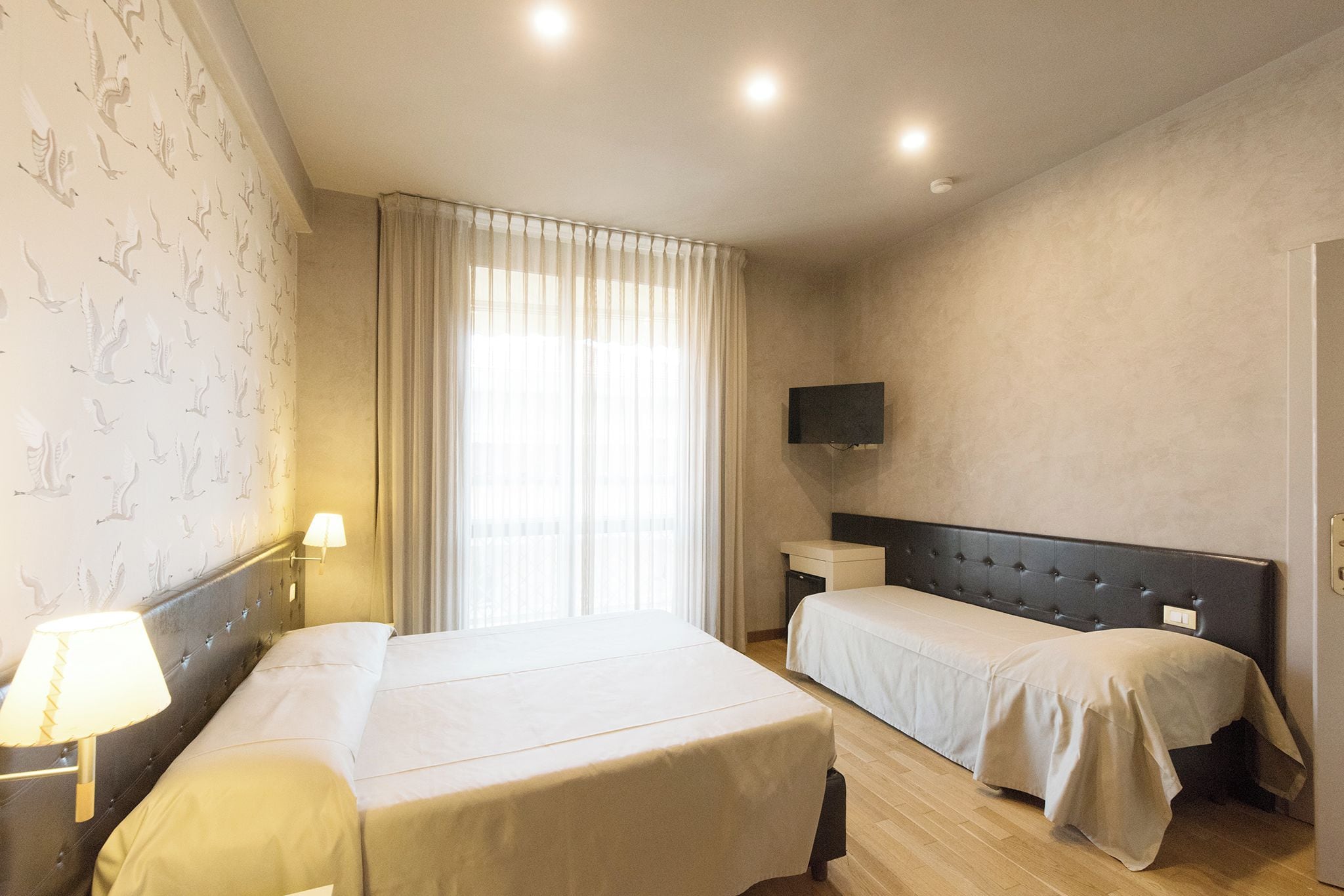 A hotel room with two beds, a flat-screen TV, a bedside table, and a window with curtains.