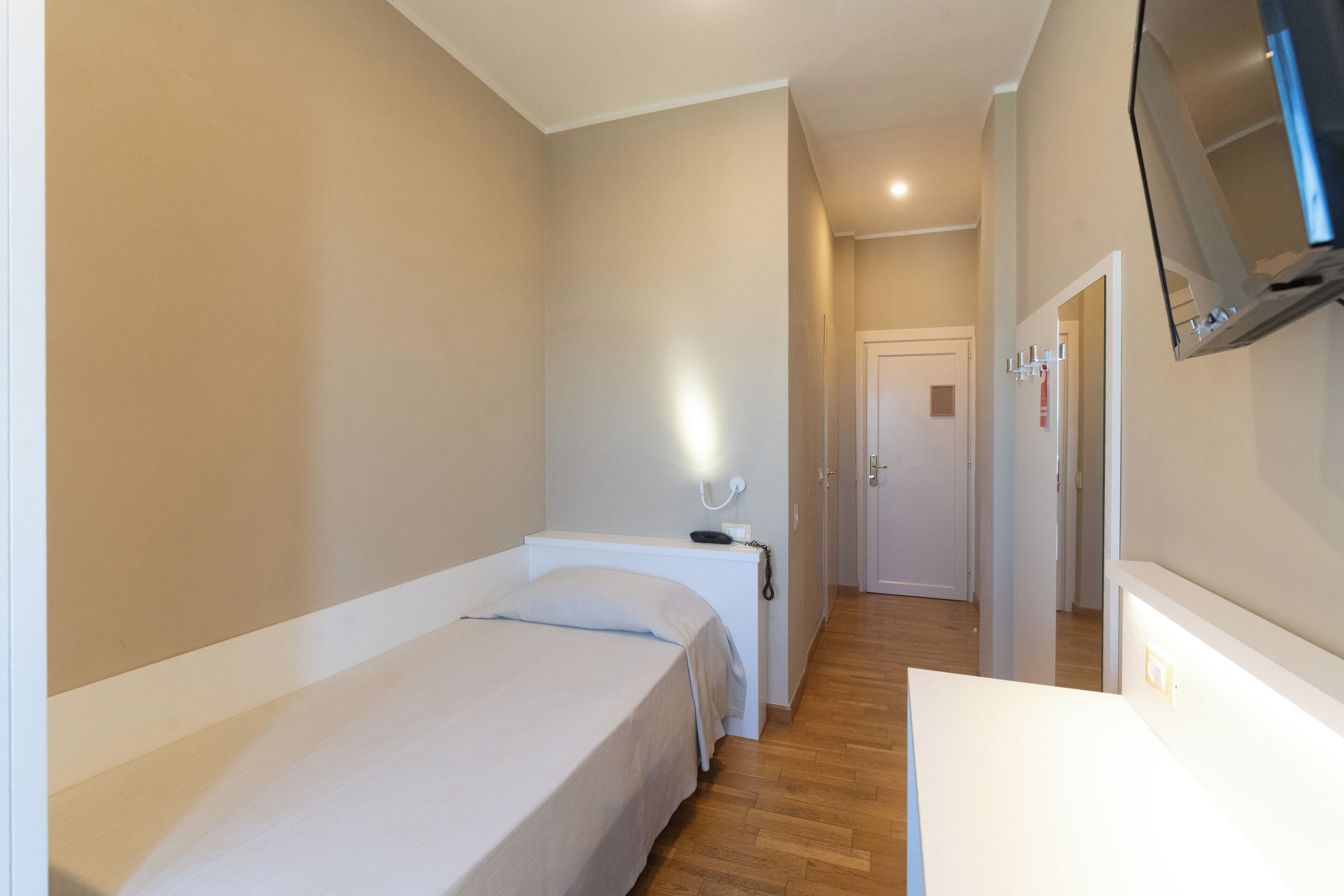 Una stanza piccola e ben illuminata, con un letto singolo, un comodino, una TV a muro e una porta che dà su un'altra stanza.