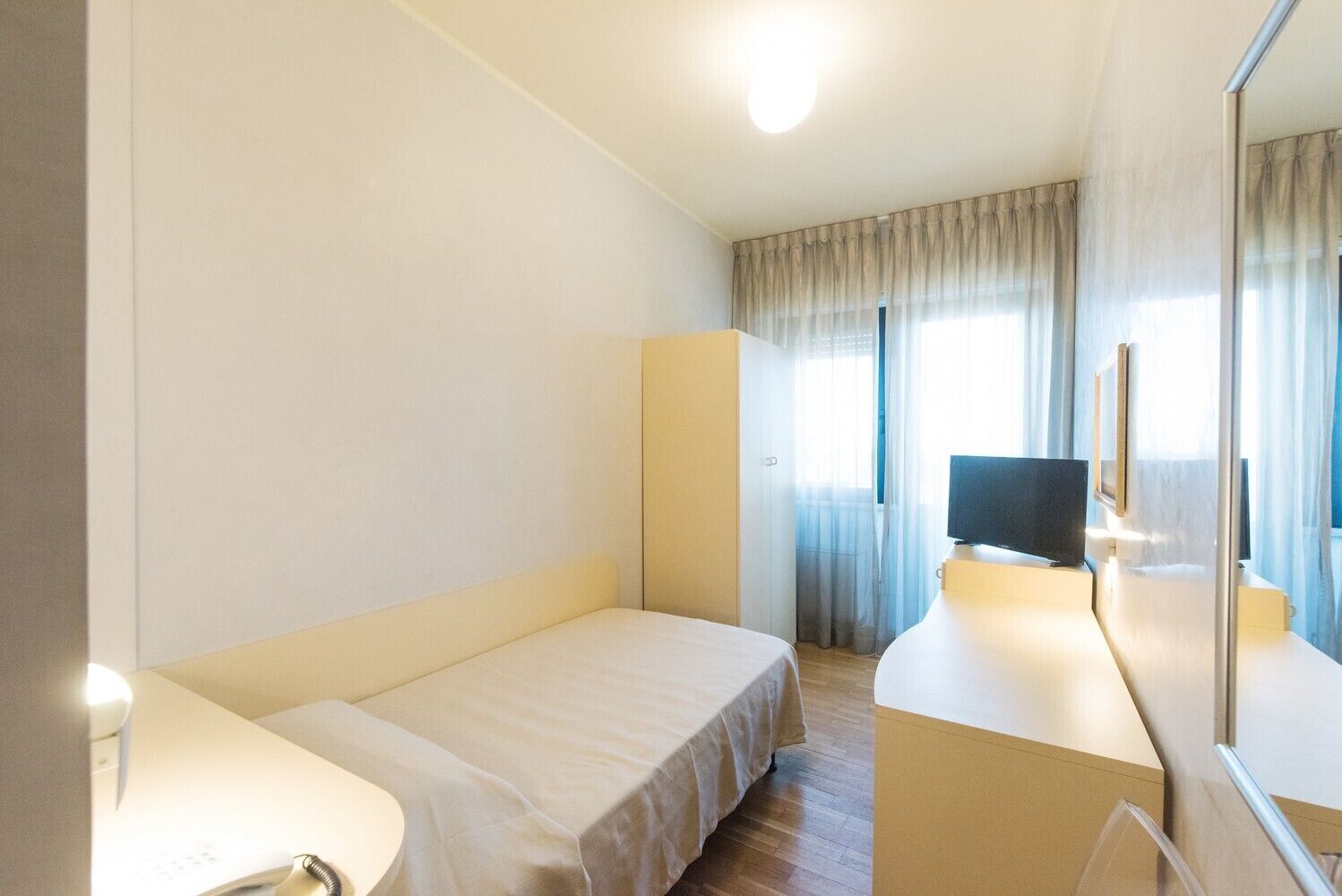 Een hotelkamer met een bed, een bureau met een computer, een spiegel en een raam met gordijnen.