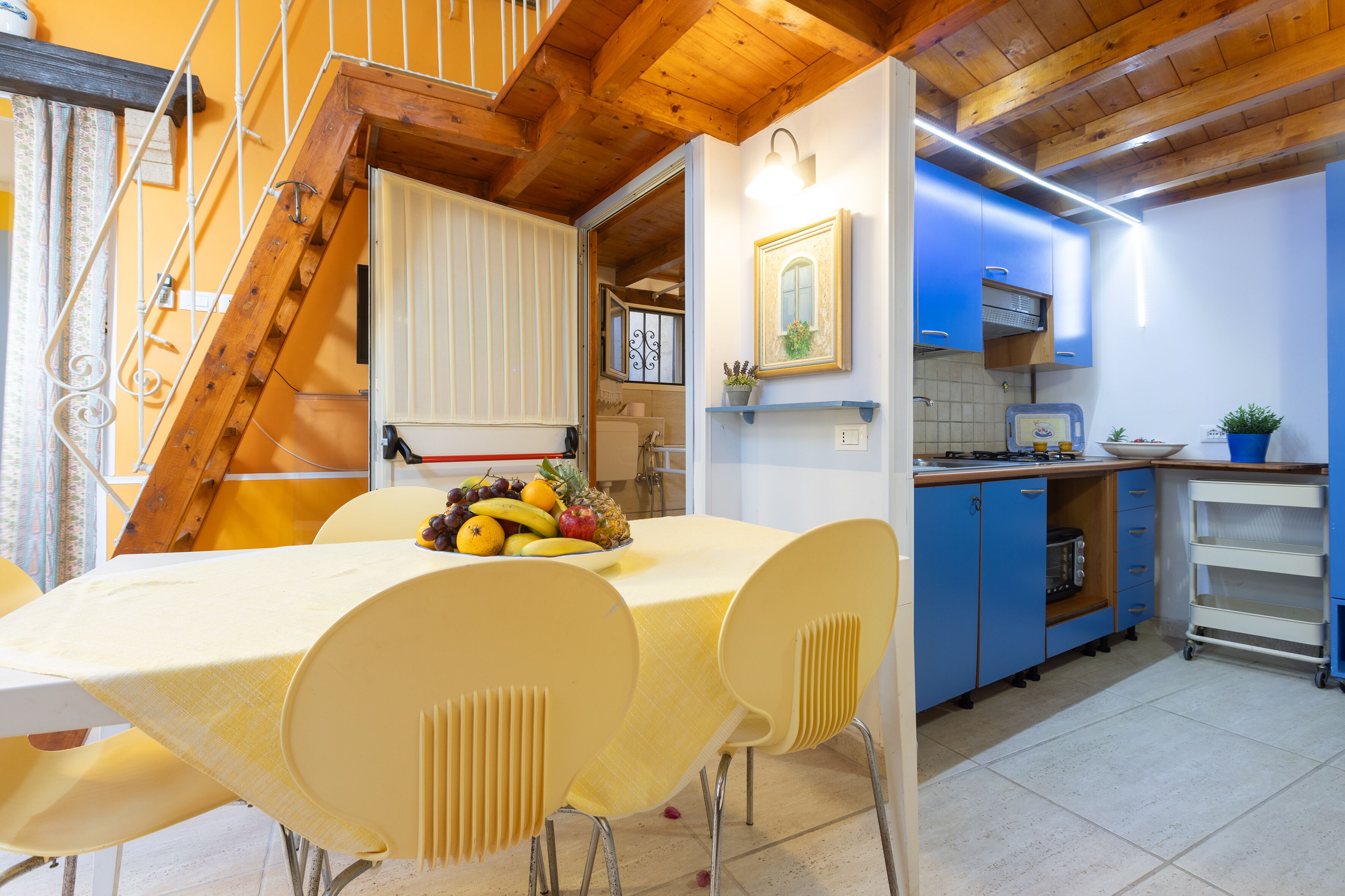 Un comedor con una mesa dispuesta para cuatro, una cocina con armarios azules y una escalera de madera.