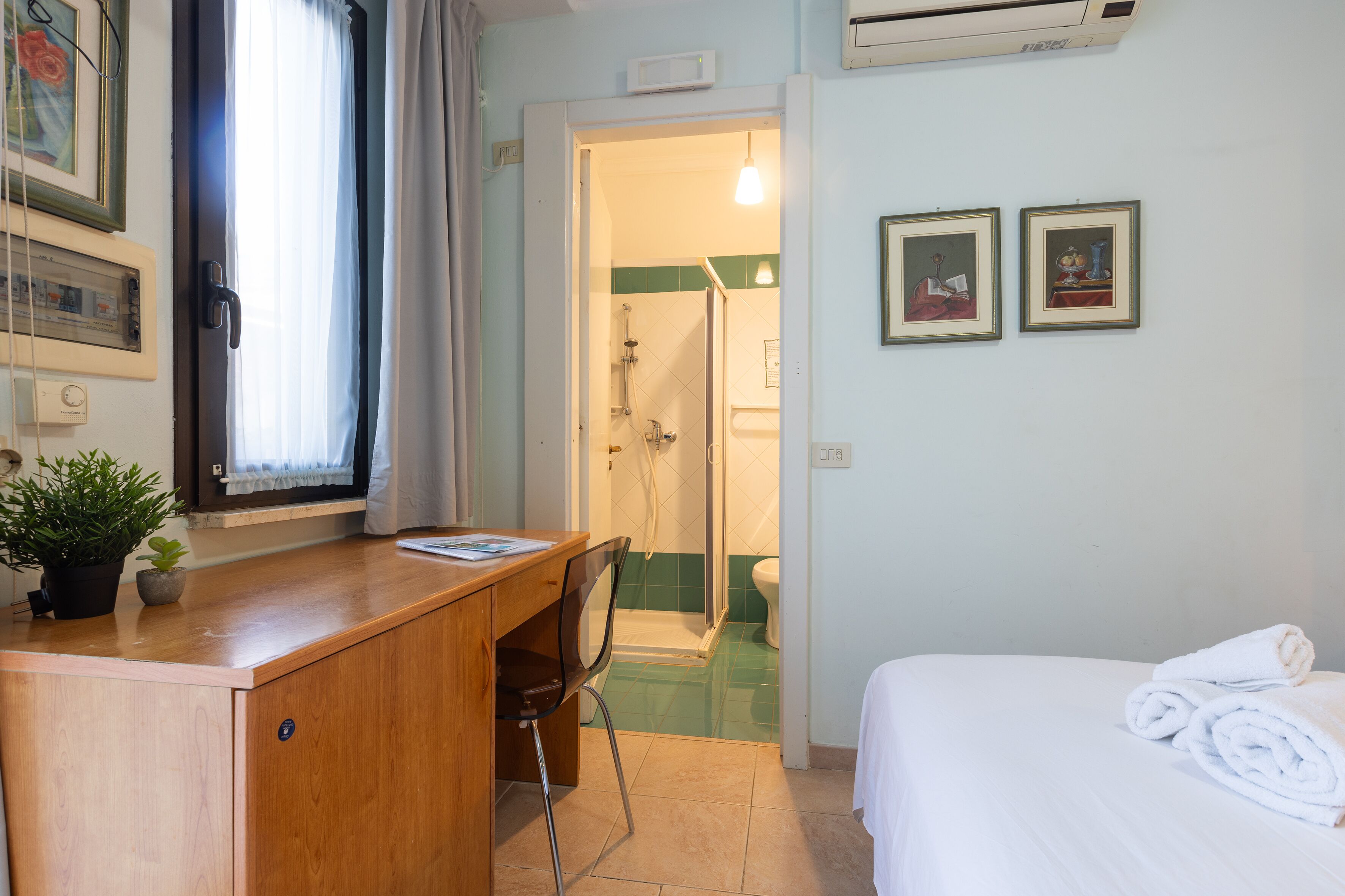 Quarto de hotel com secretária de madeira, uma cadeira, cama com roupa branca, área de chuveiro e duas molduras na parede.