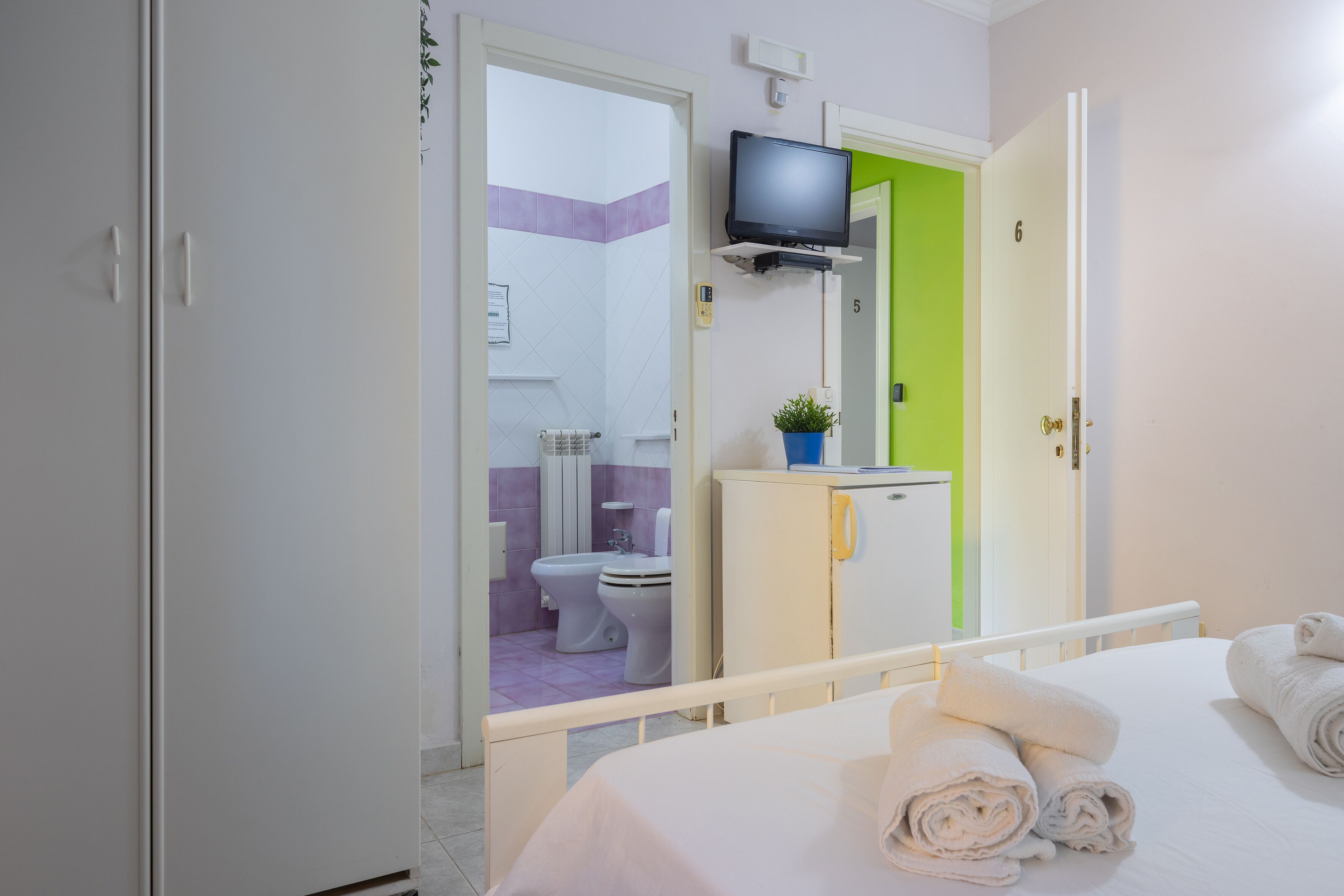 A hotel room with a bed, a TV mounted on the wall, a bathroom visible through an open door, and a small cabinet with a plant.