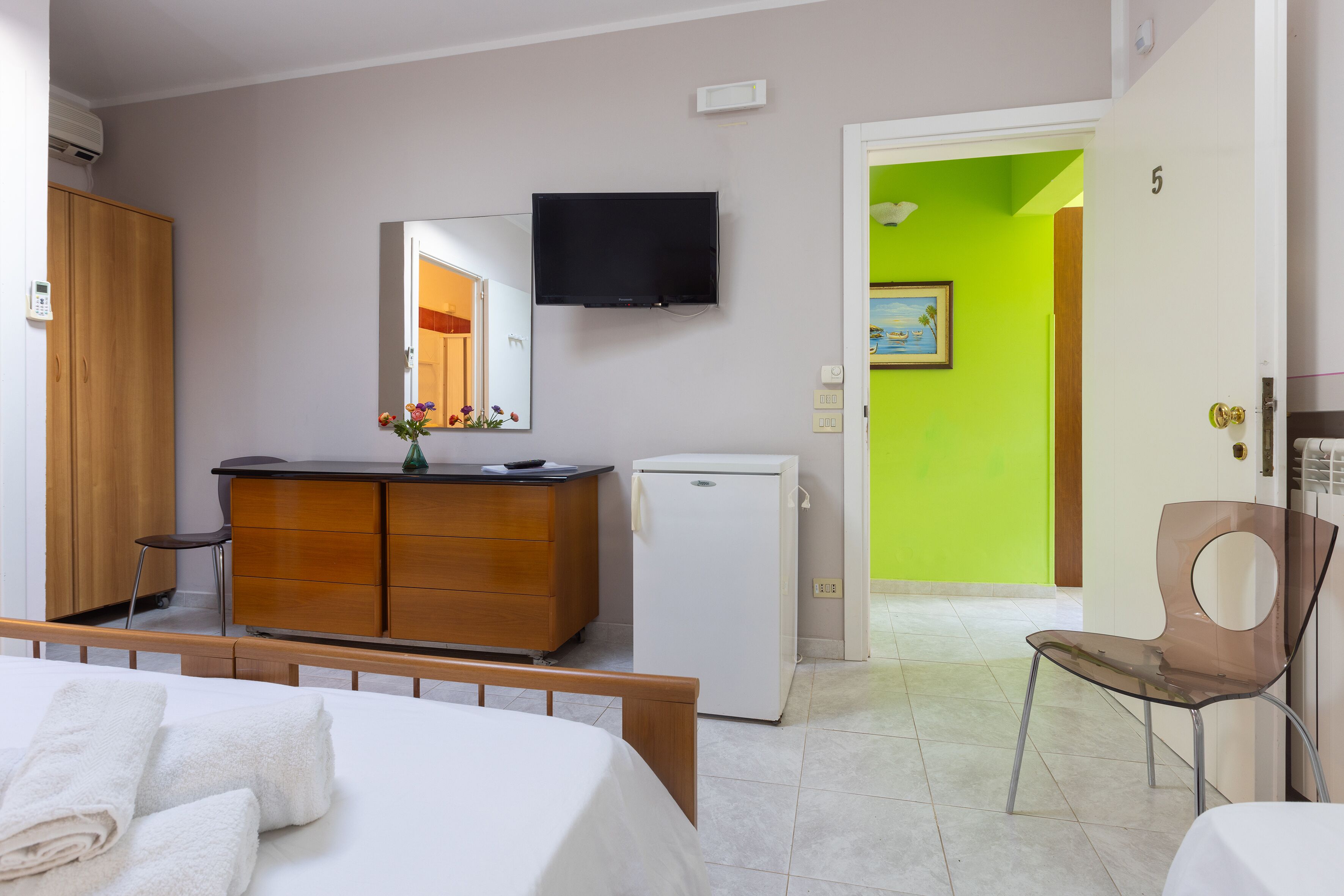 Een hotelkamer met een bed, een houten dressoir, een witte koelkast, een aan de muur bevestigde televisie en een groene wand in de aangrenzende kamer.