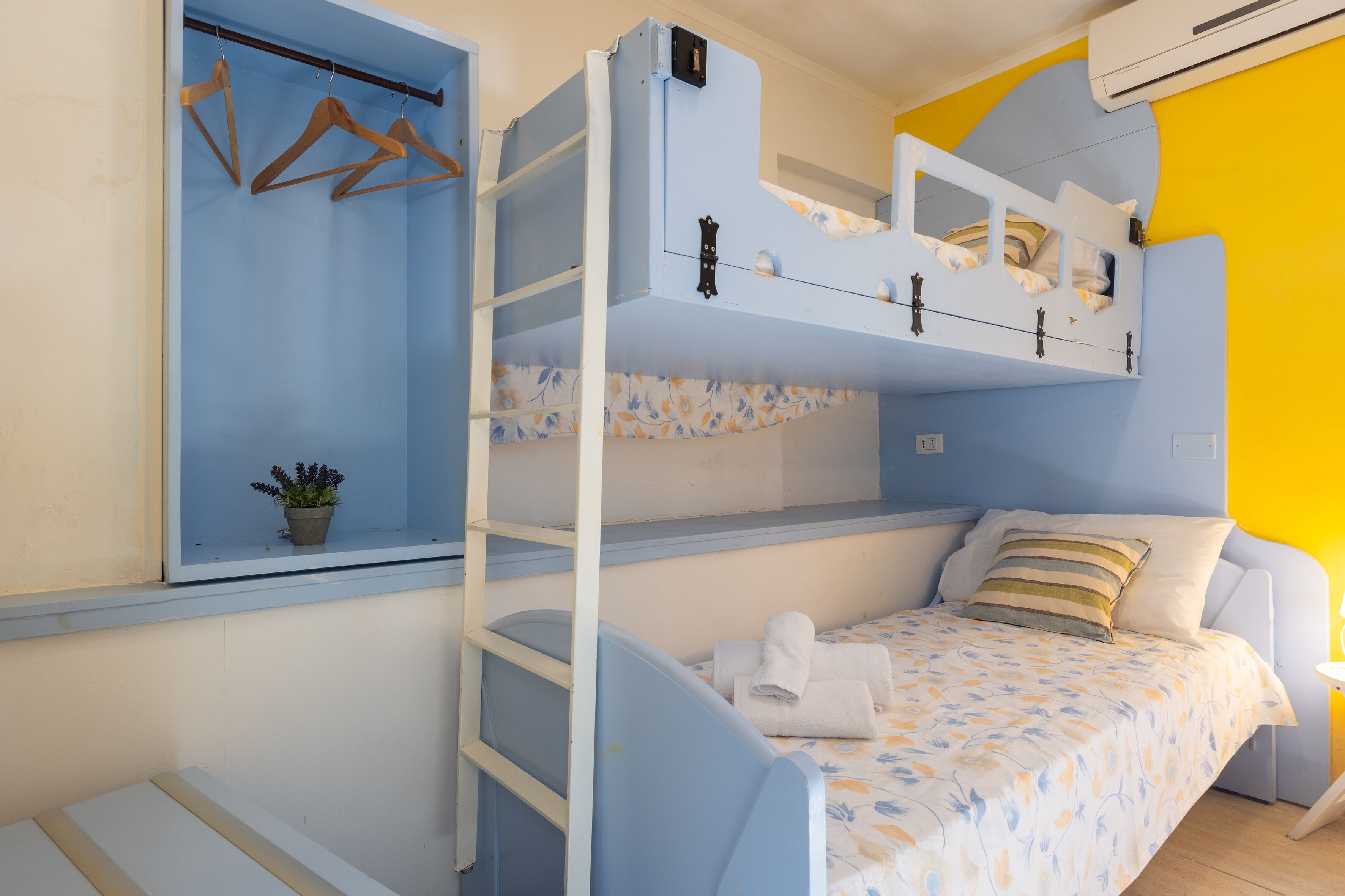 A bunk bed room with a window, a shelf with a potted plant, and a bedside table with a lamp.