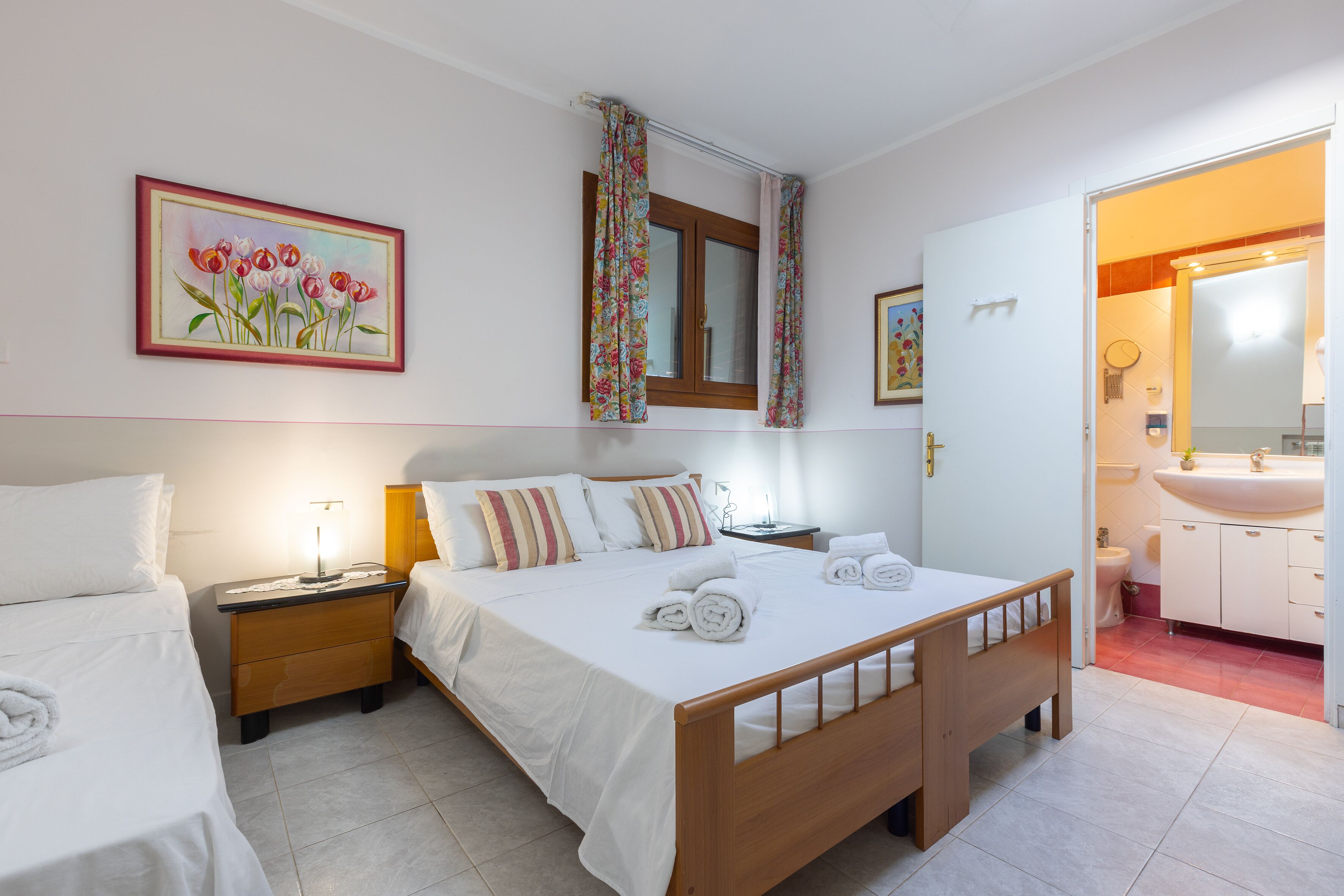 A hotel room with a wooden bed, two bedside tables, a nightstand, a painting of flowers, a window with curtains, and a bathroom visible through an open door.