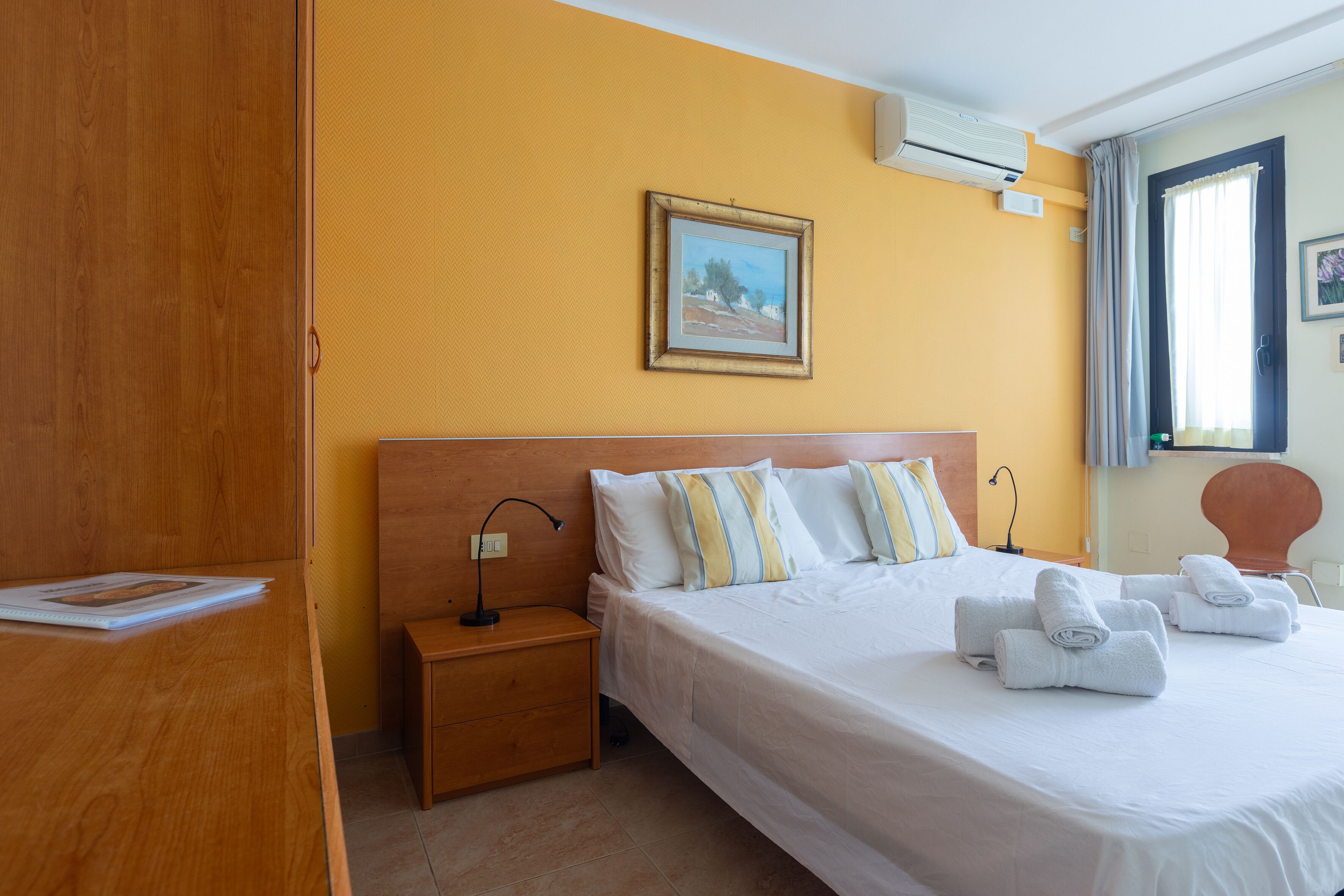 A hotel room with a wooden wardrobe, a bed with white bedding and striped pillows, a nightstand with a lamp, and a painting on the wall.