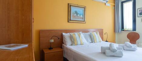 A hotel room with a wooden wardrobe, a bed with white bedding and striped pillows, a nightstand with a lamp, and a painting on the wall.