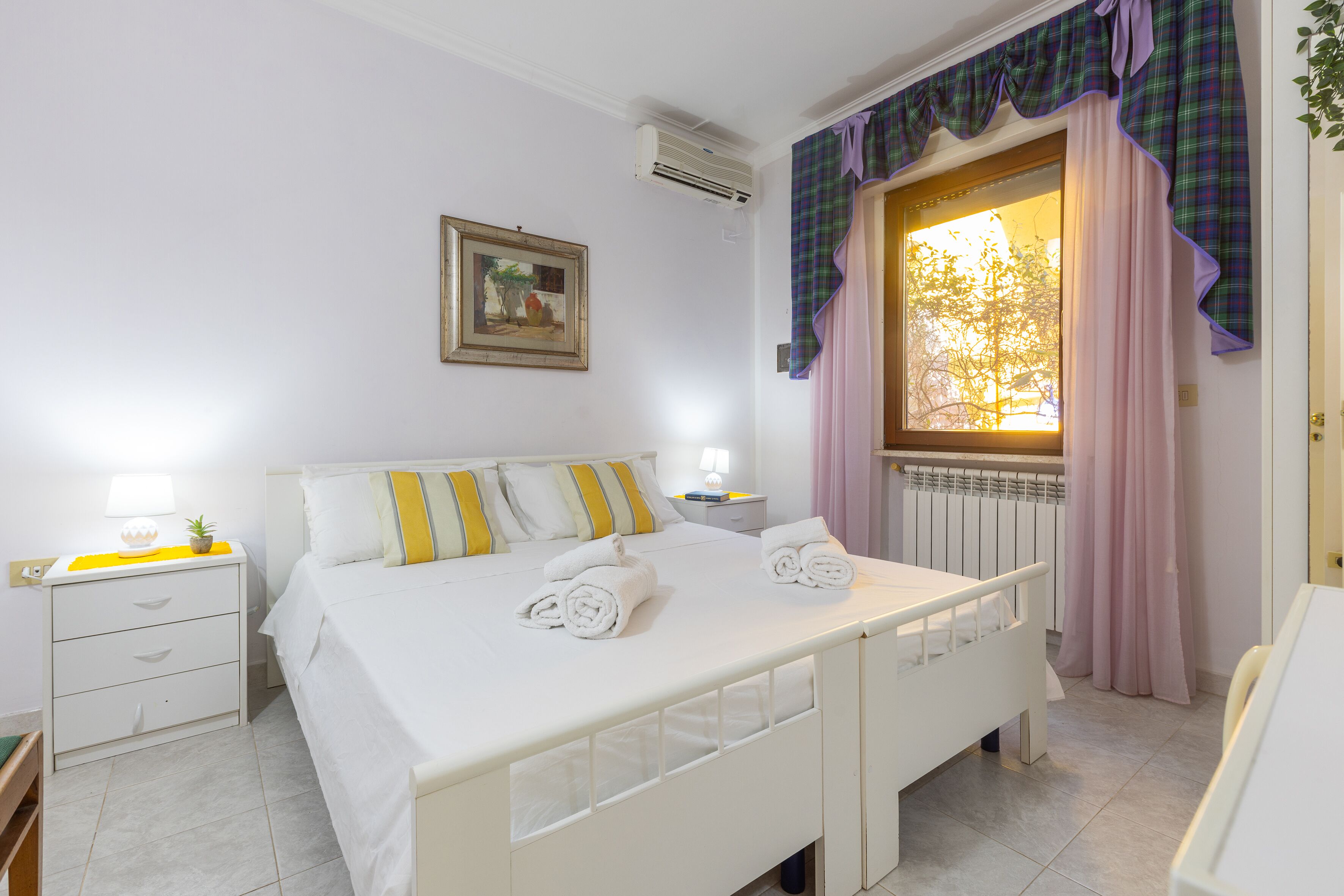 Une chambre à coucher comprenant un lit, des tables de chevet, une commode, une fenêtre avec des rideaux et un radiateur.