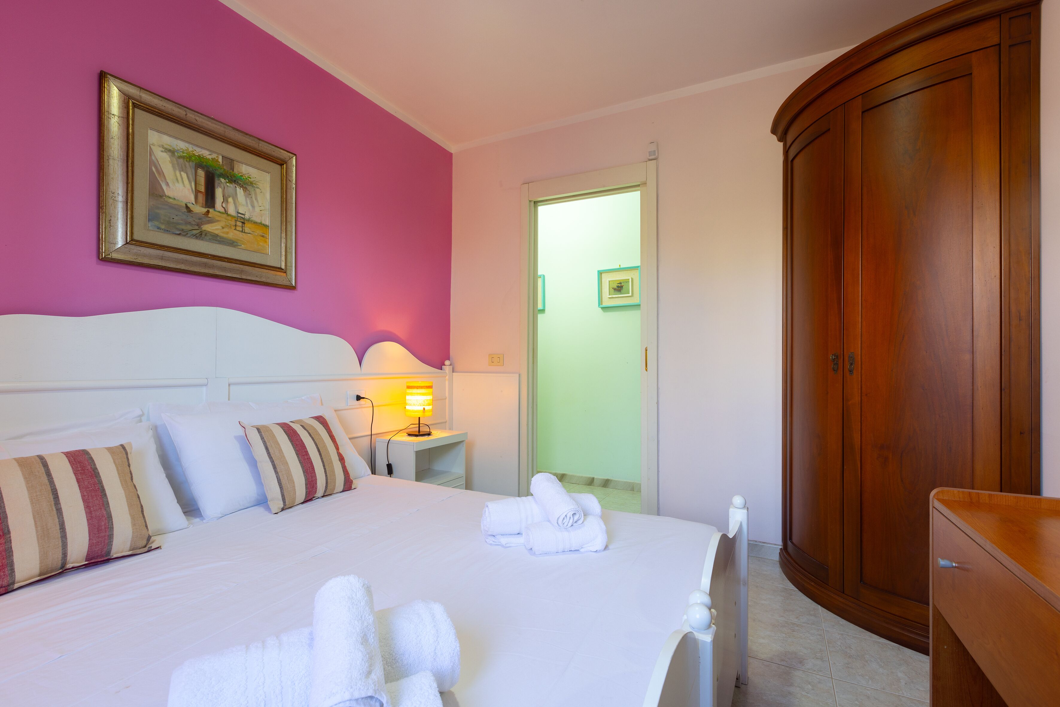 A hotel room with a bed, pillows, a bedside table with a lamp, a wardrobe, and a door leading to another room.