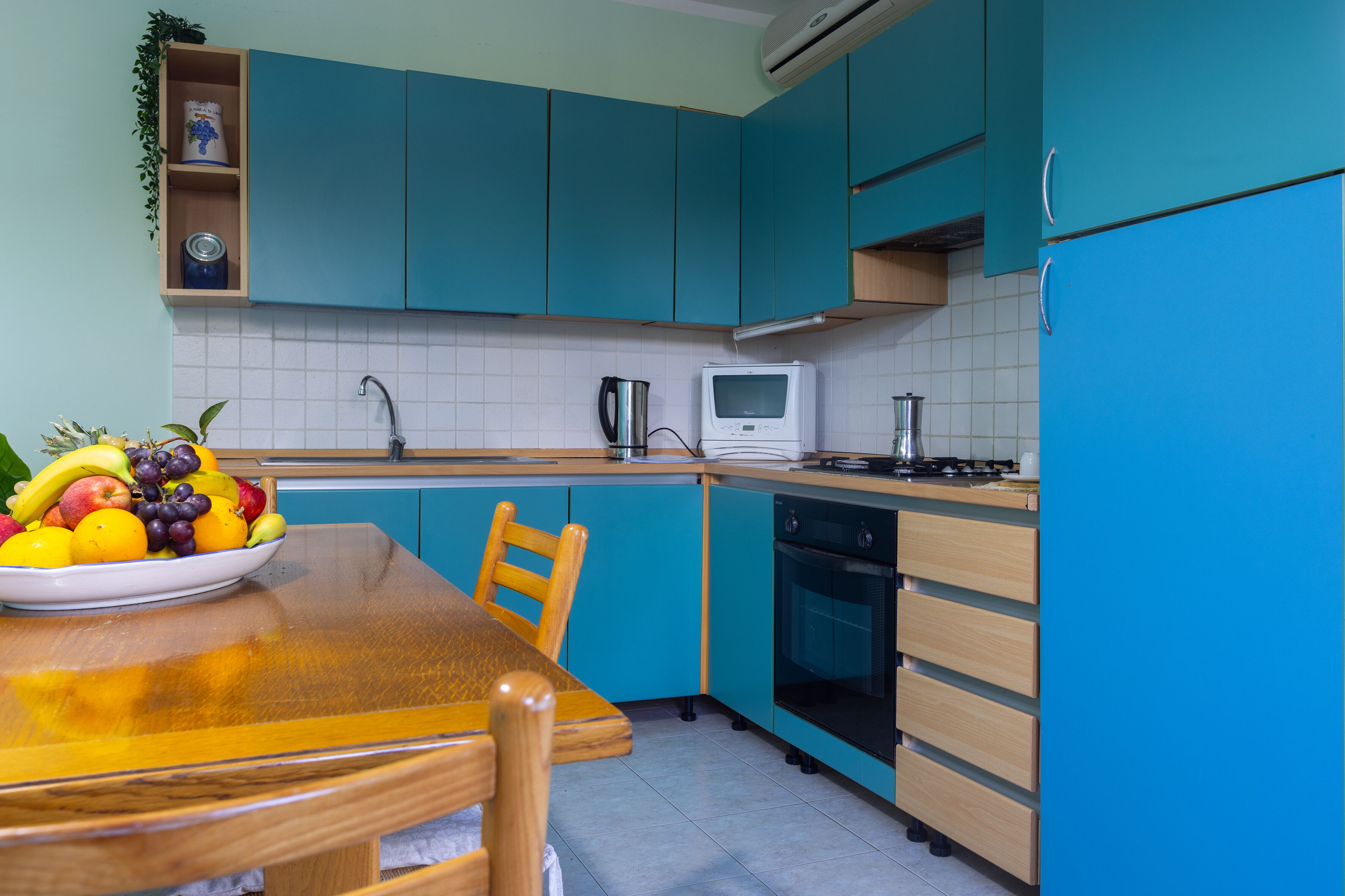 Una cocina con armarios verde azulado, una mesa de madera con un frutero, un microondas y un horno.
