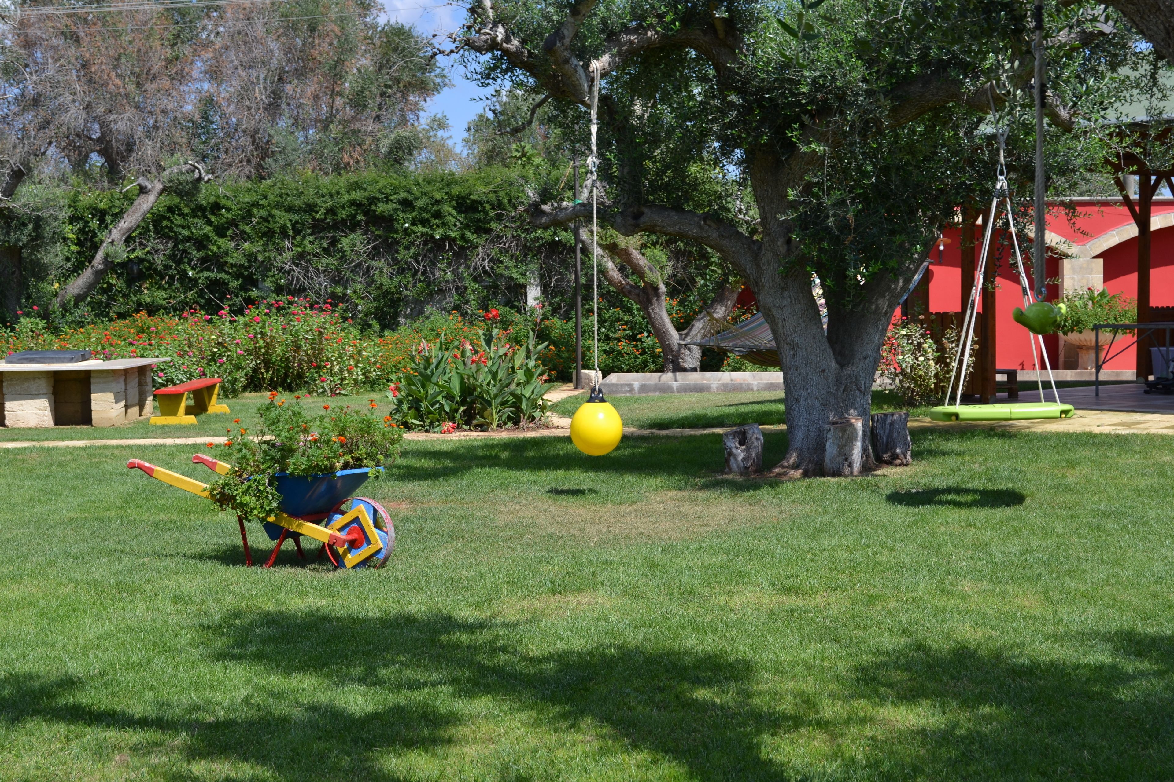 Um jardim bem cuidado com balanço, uma bola amarela e um carrinho de mão com plantas.