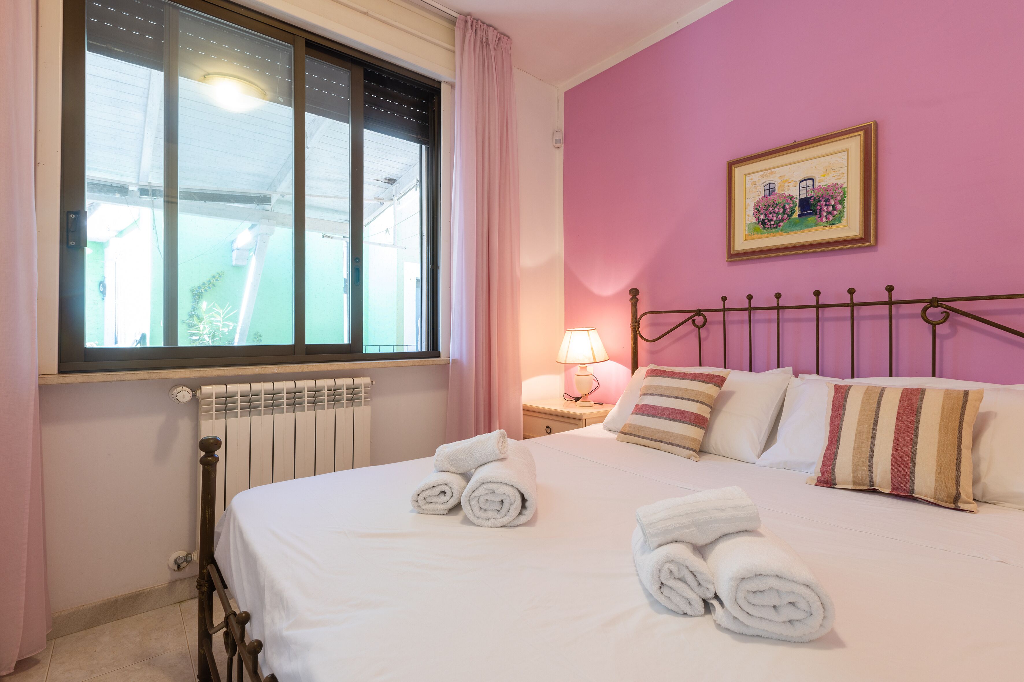 A hotel room with a bed, pillows, a window, a radiator, and a lamp.