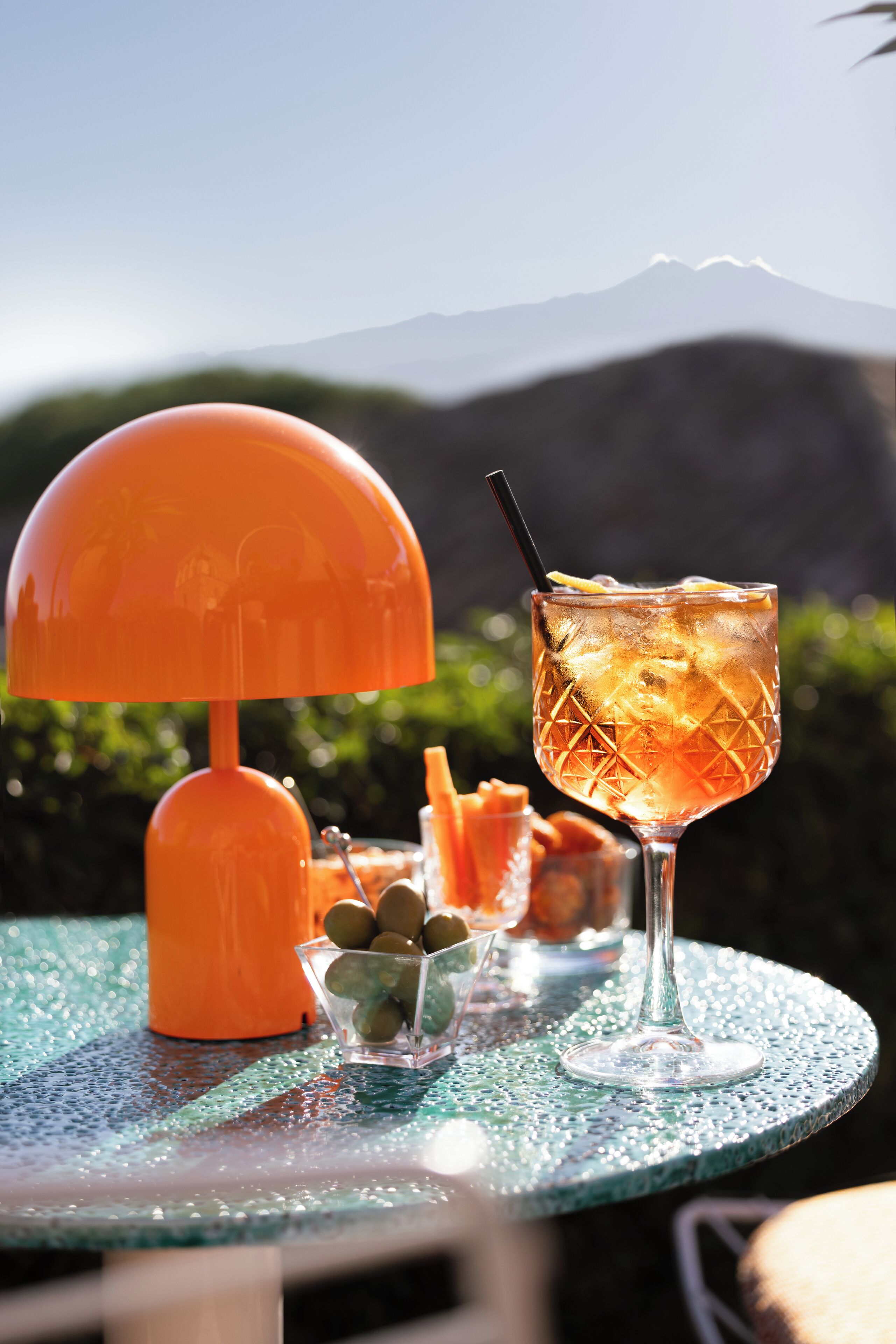 Una mesa con un vaso de bebida helada, un bol de aceitunas y una lámpara naranja.