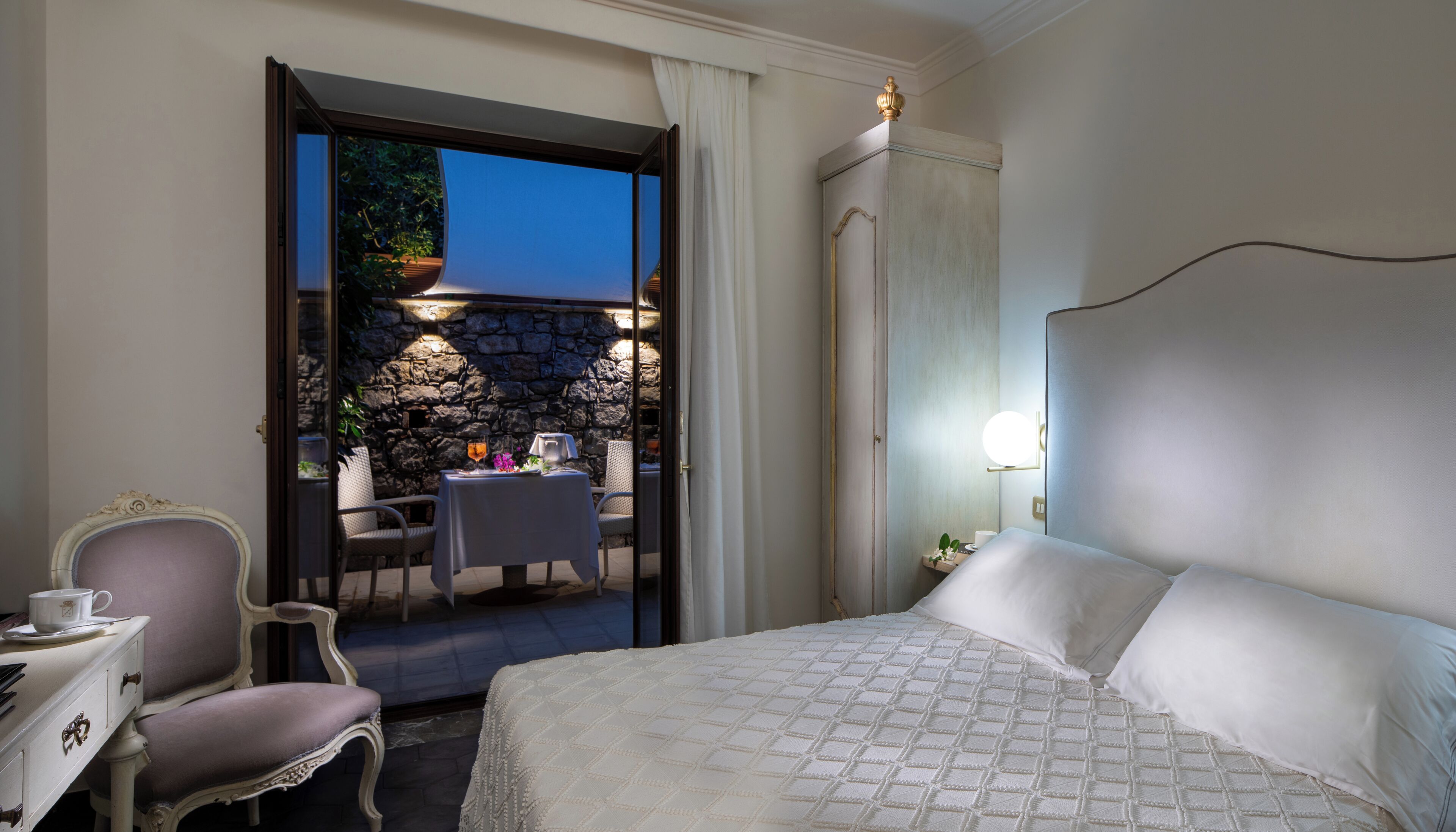 A hotel room with a bed, a table with chairs, a small balcony with a stone wall, and a view of the outdoors.