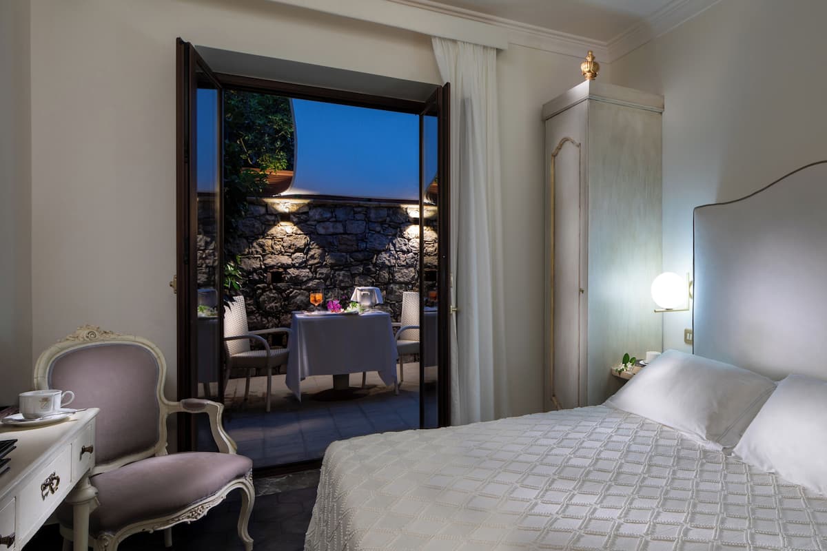 A hotel room with a bed, a table with chairs, a small balcony with a stone wall, and a view of the outdoors.
