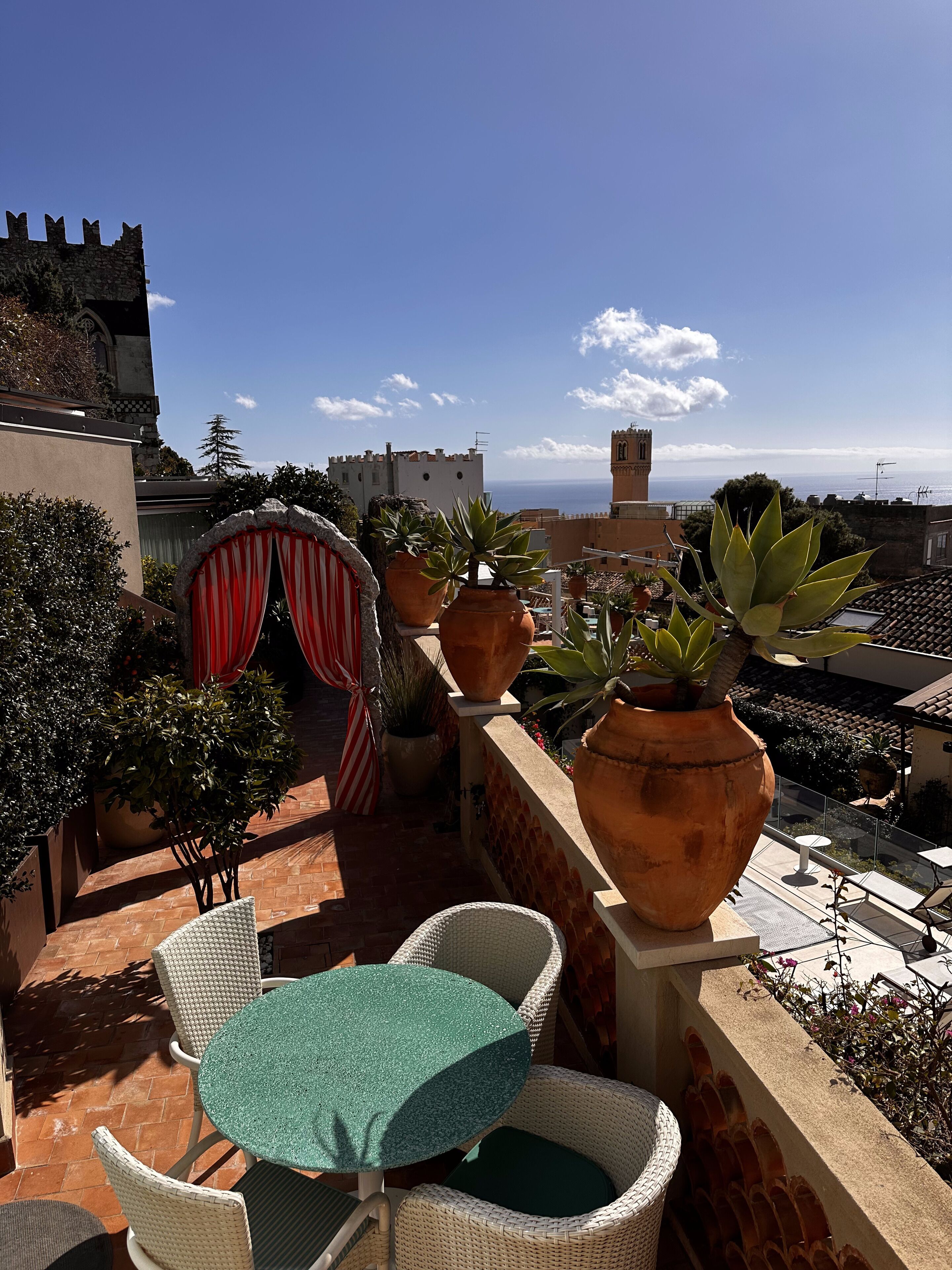 Terrassa al terrat amb cadires de vimet, una taula, plantes en testos i vista al mar i edificis històrics.