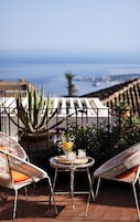 A terrace with two wicker chairs, a small round table with a drink, and a view of the sea.