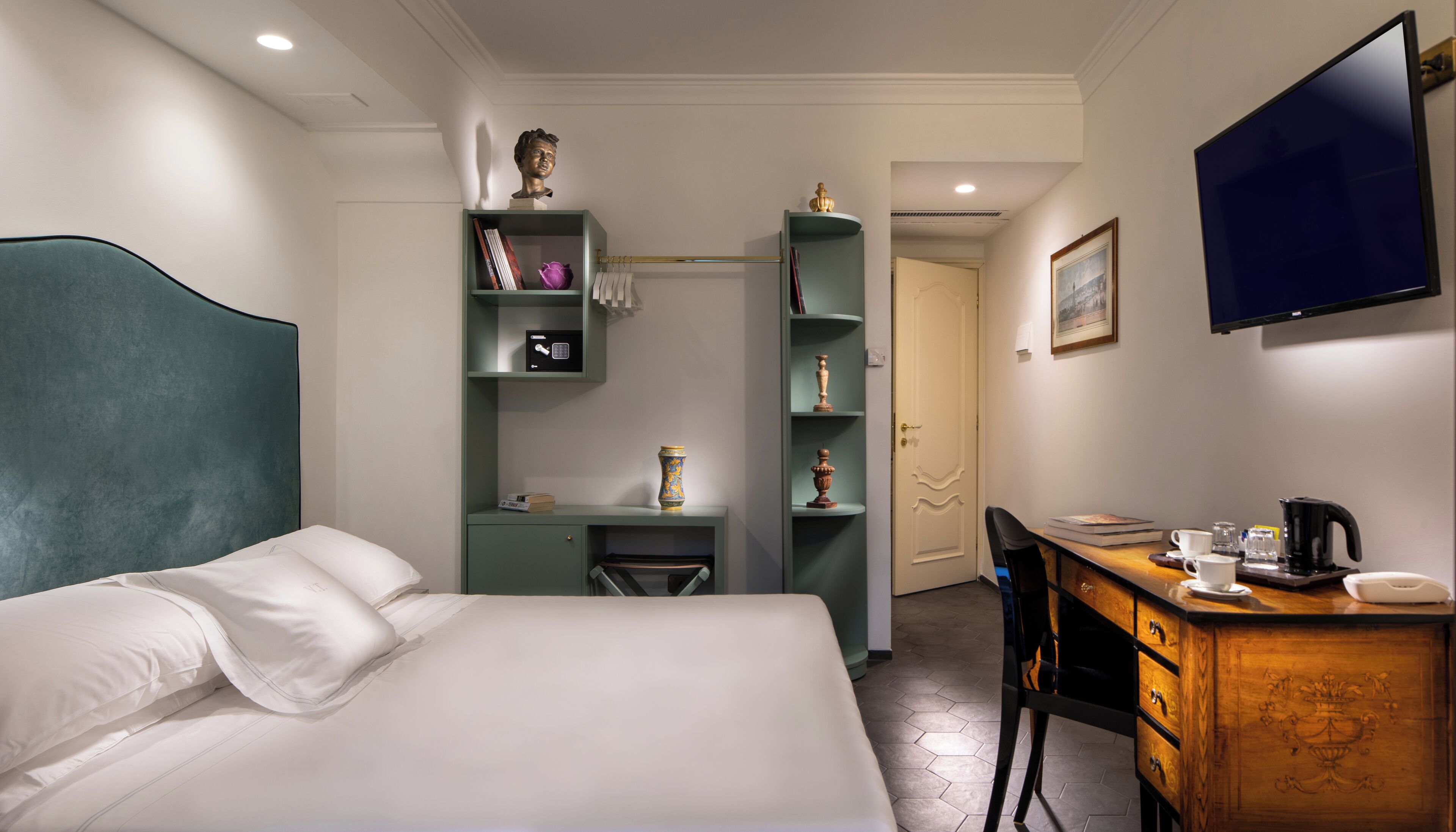 A hotel room with a bed, a desk, a chair, a TV, and shelves with decorative items.
