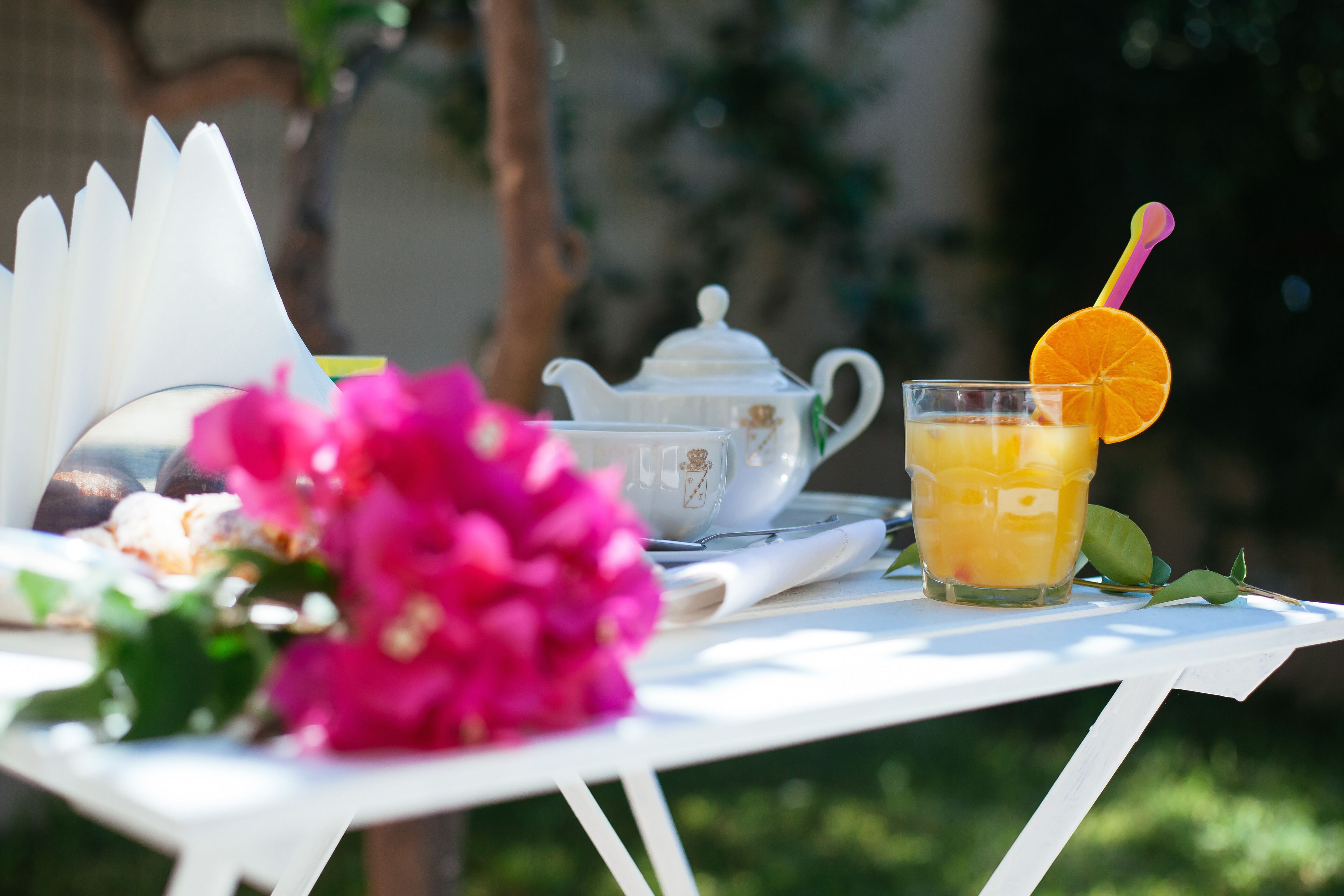 Una mesa blanca con un calentador de agua, un vaso de jugo de naranja y una flor rosa.