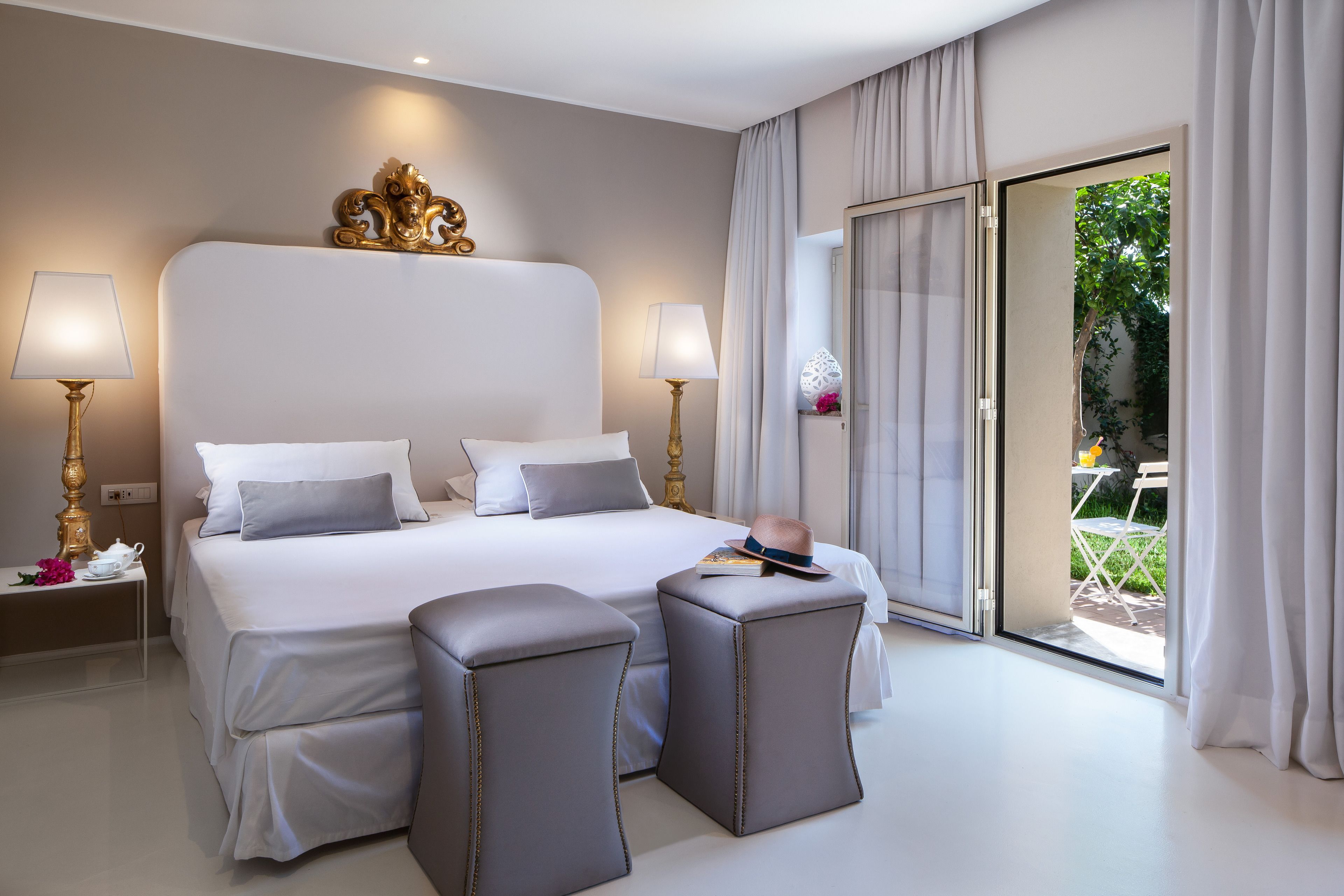 Ein modernes Hotelzimmer mit einem großen Bett, zwei Nachttischen, einer Sitzecke mit Poufs und einer Glastür zu einem Außenbereich.