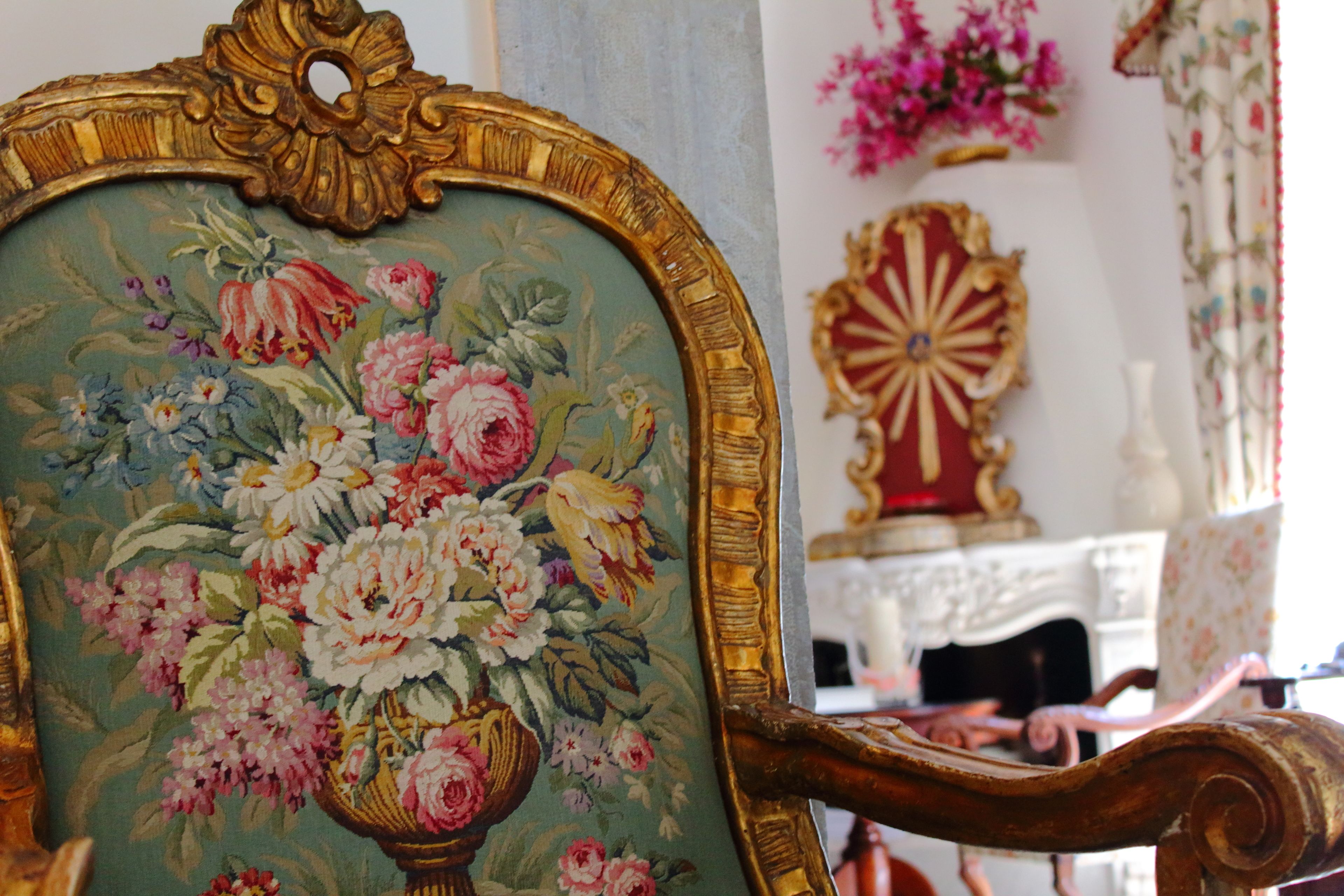 A vintage chair with a floral tapestry, a decorative mirror, and a floral arrangement on the wall.