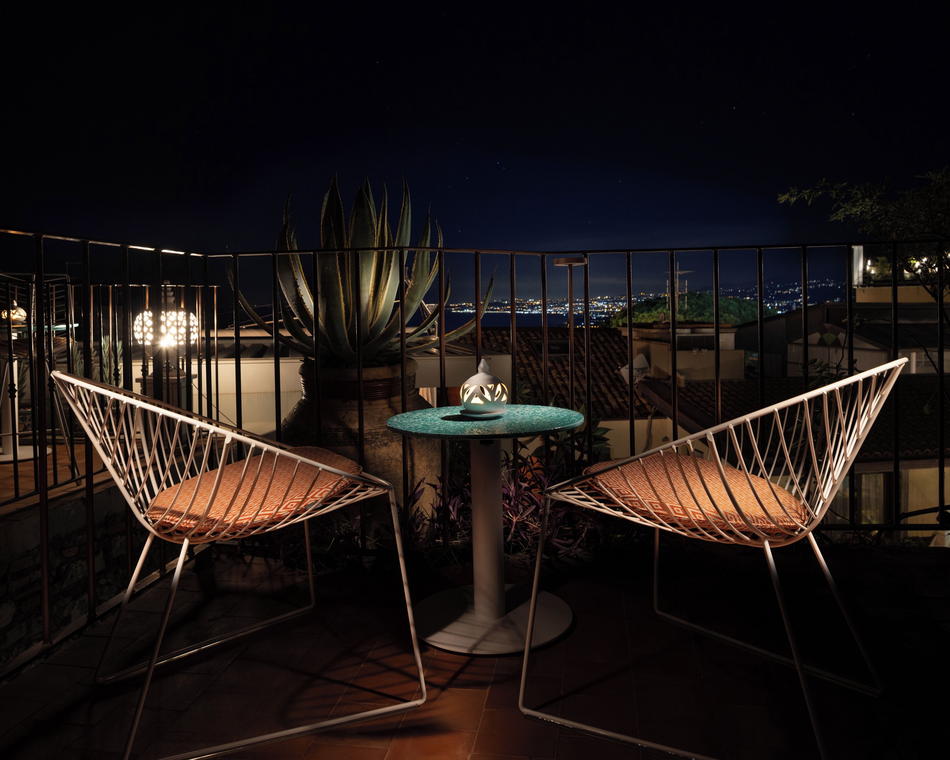 A balcony with two wireframe chairs and a small table, surrounded by a metal railing.