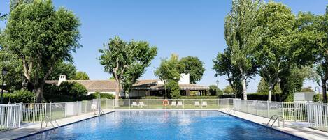 A swimming pool with white railings, surrounded by trees and a building with a tiled roof in the background.